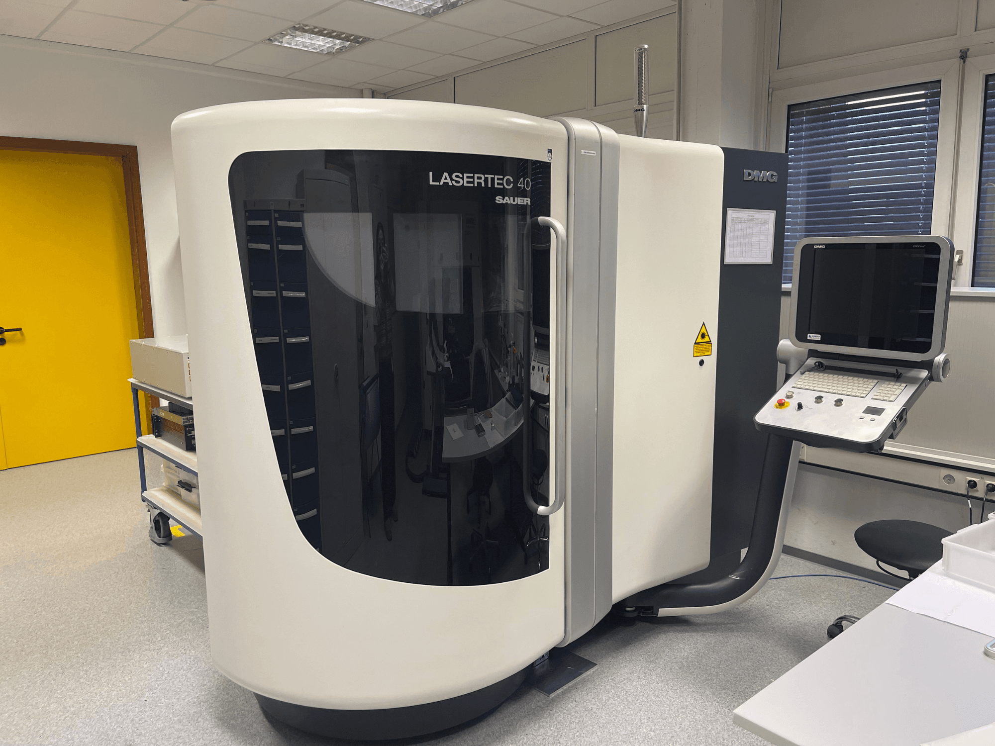 DMG MORIn valkoinen LASERTEC 40 -jyrsinkone, etukuva, jossa on ohjauspaneeli ja ergonominen muotoilu hyvin valaistussa huoneessa.