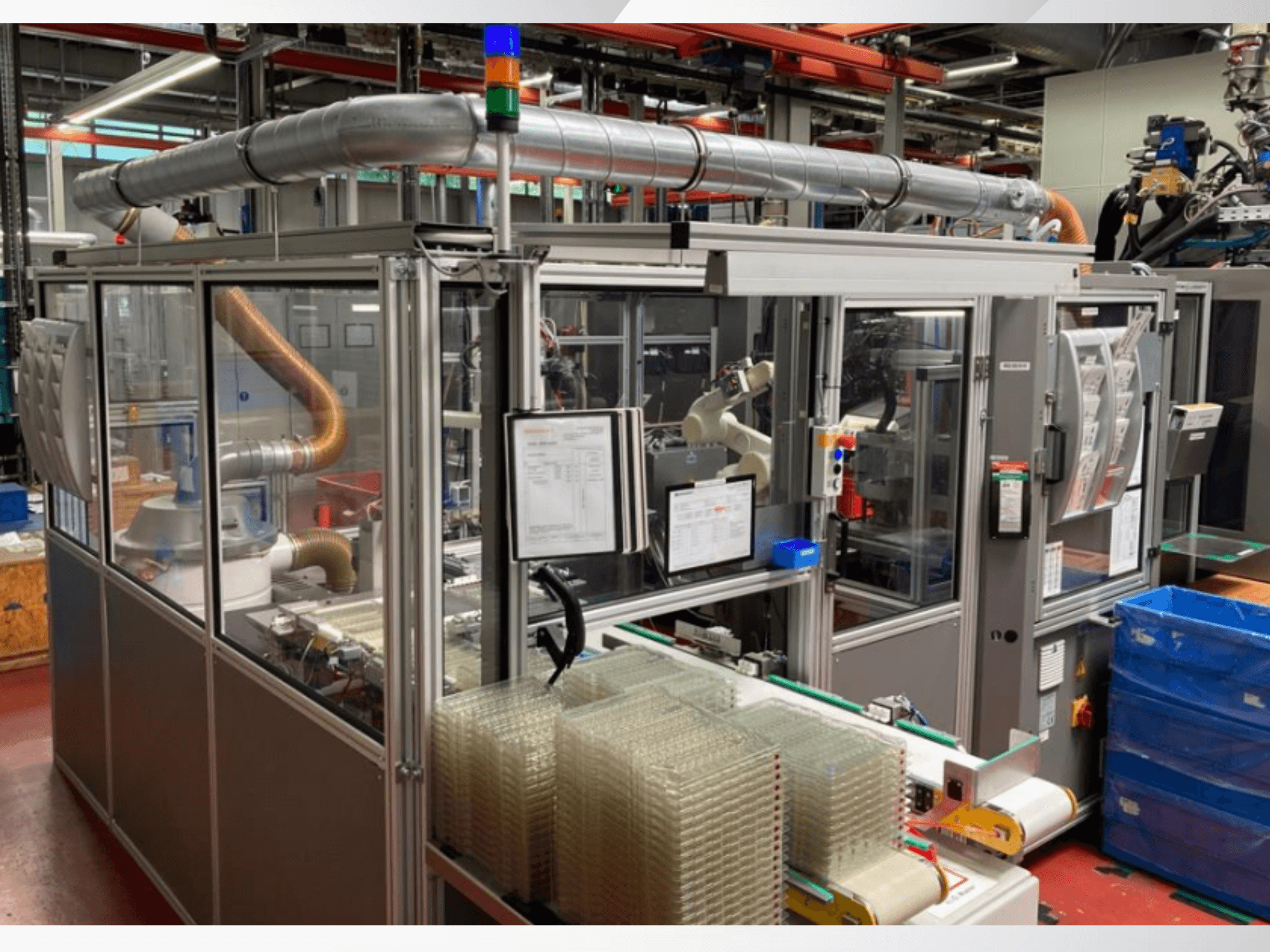 Automaattinen kokoonpanokone, jossa on robottivarsi, läpinäkyvät sivupaneelit ja pinotut kirkkaiden tuotteiden säiliöt, etukuva.