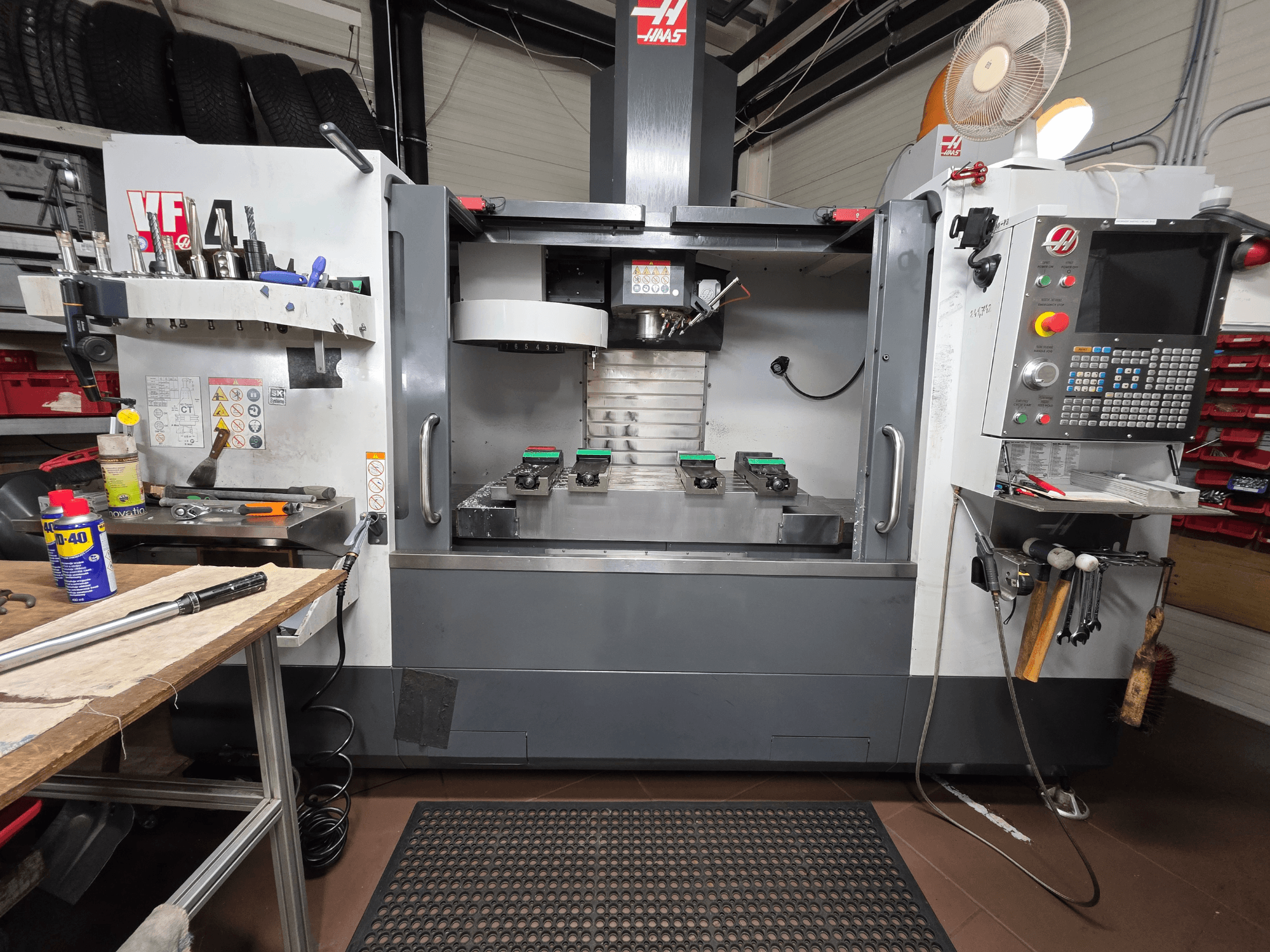 Haas VF-4 CNC-jyrsimen etukuva työpajassa, jossa on työkaluja, tietokoneen käyttöliittymä ja kaksi työkappaletta pöydällä.