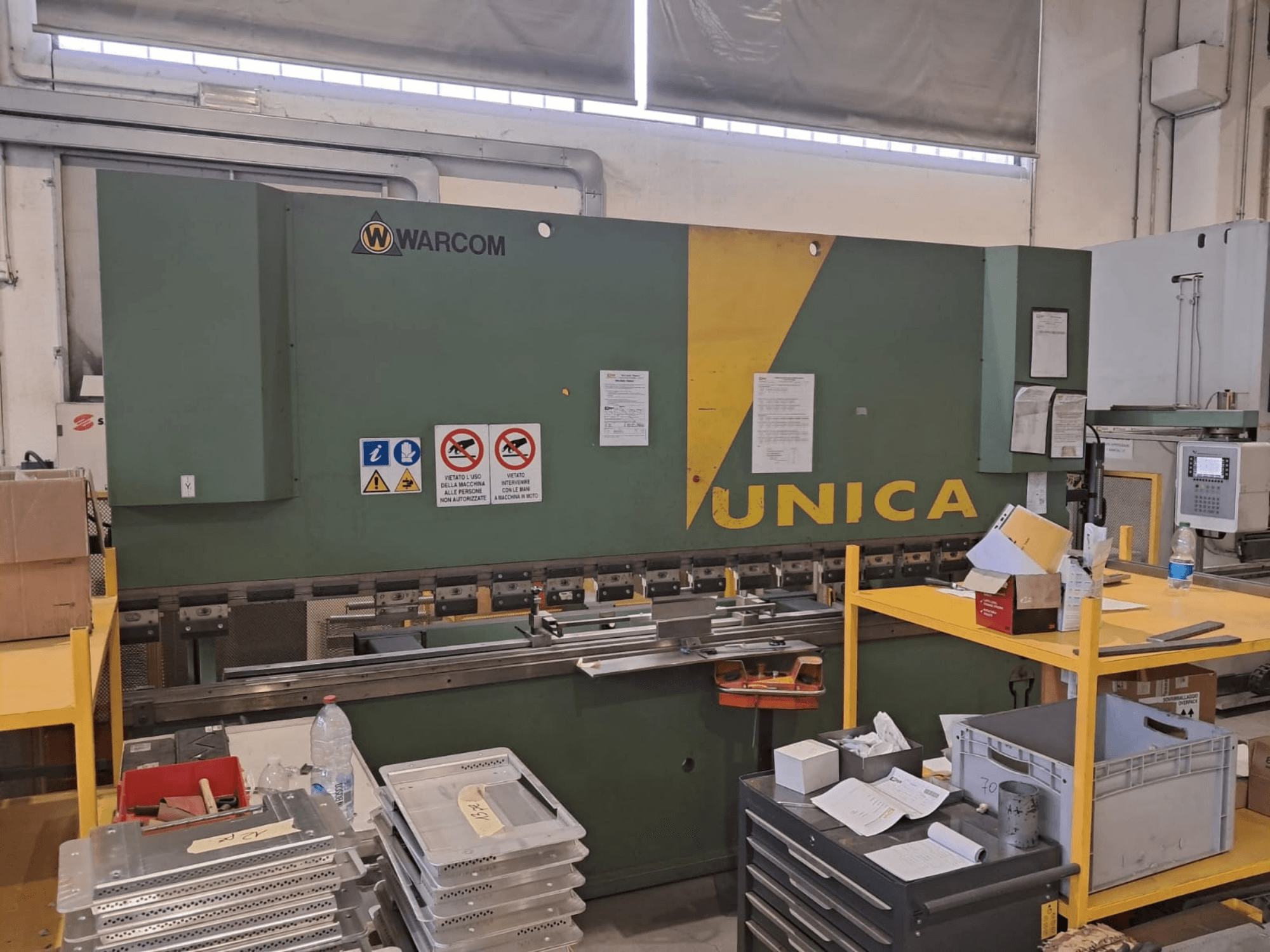 Warcom UNICA 100-40 -hydraulipuristin, etunäkymä, vihreä runko keltaisilla korostuksilla, turvallisuusmerkit näkyvissä, työkalut ja materiaalit pöydällä.