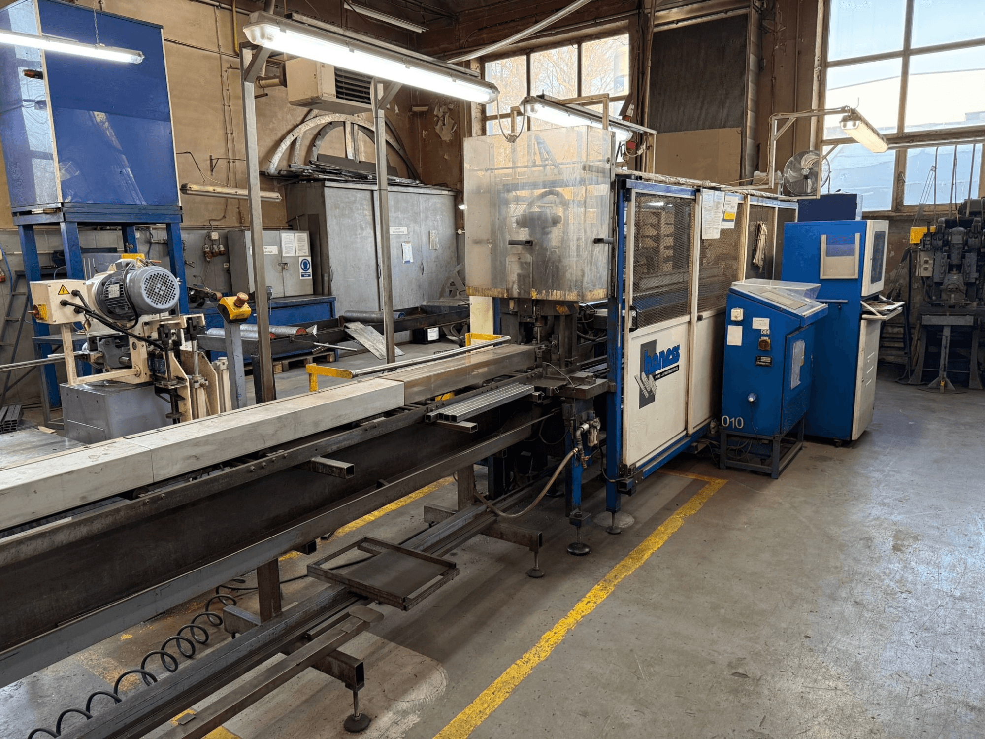 Teollisuusverstas, jossa on sininen Haweas-kone, sivukuva, jossa näkyy kuljetin ja laitteet suuressa, hyvin valaistussa tilassa.