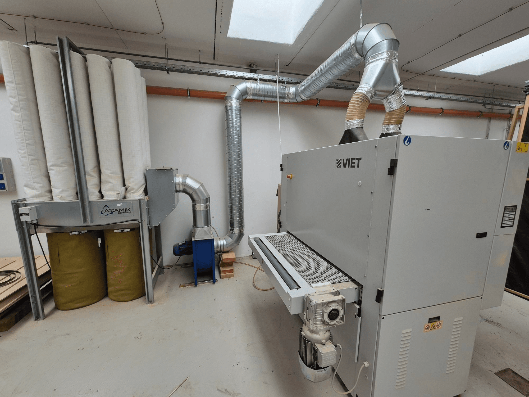 Viet pölynpoistojärjestelmä, jossa on sylinterimäiset suodattimet ja hopeanvärinen pakokaasu, edestä katsottuna työpajassa.