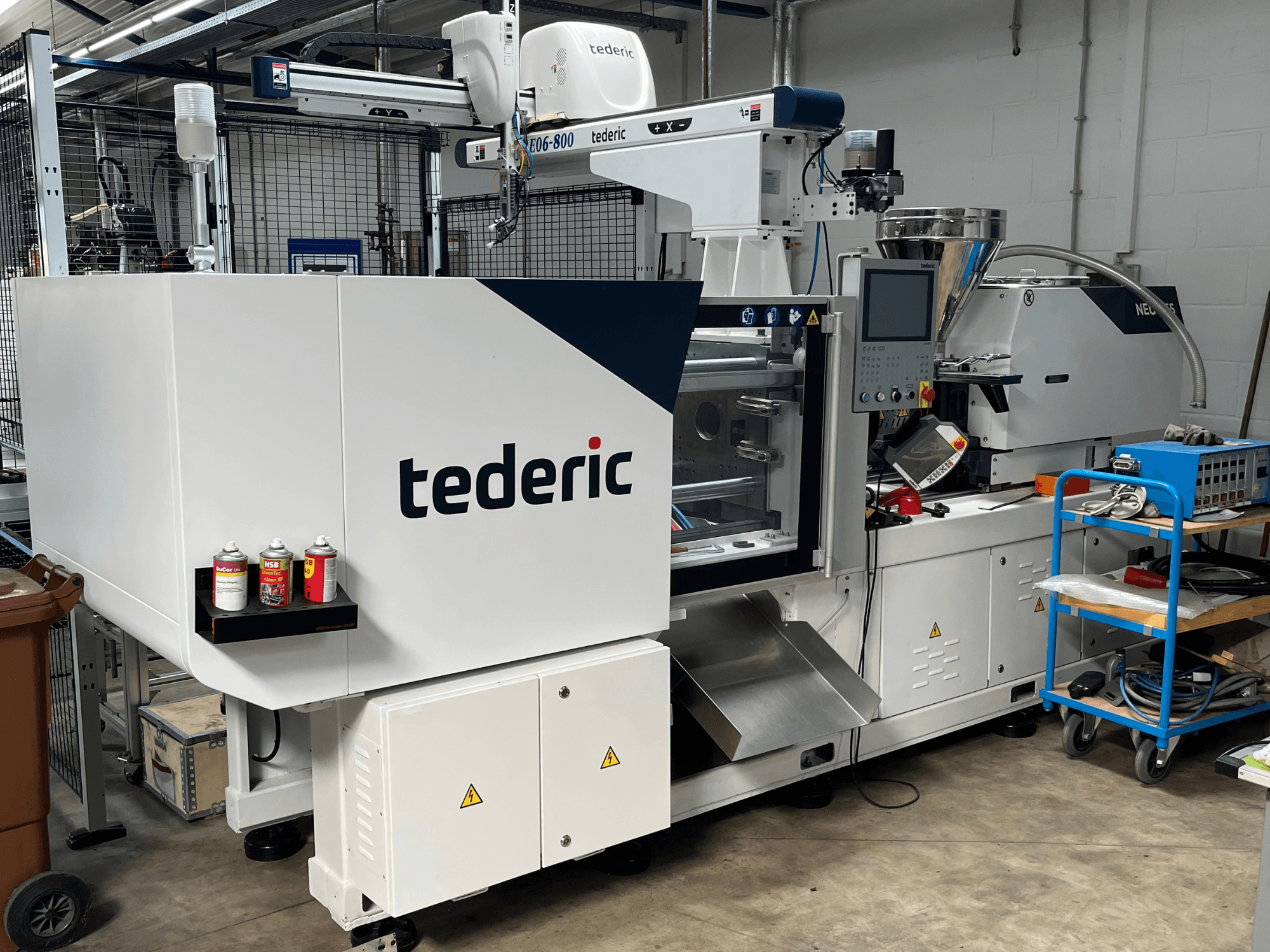 Hydraulinen ruiskuvalukone TEDERIC NEO.E55/e110h työpajassa, sivukuva, jossa näkyy ohjauspaneeli ja sen vieressä olevat työkalut.