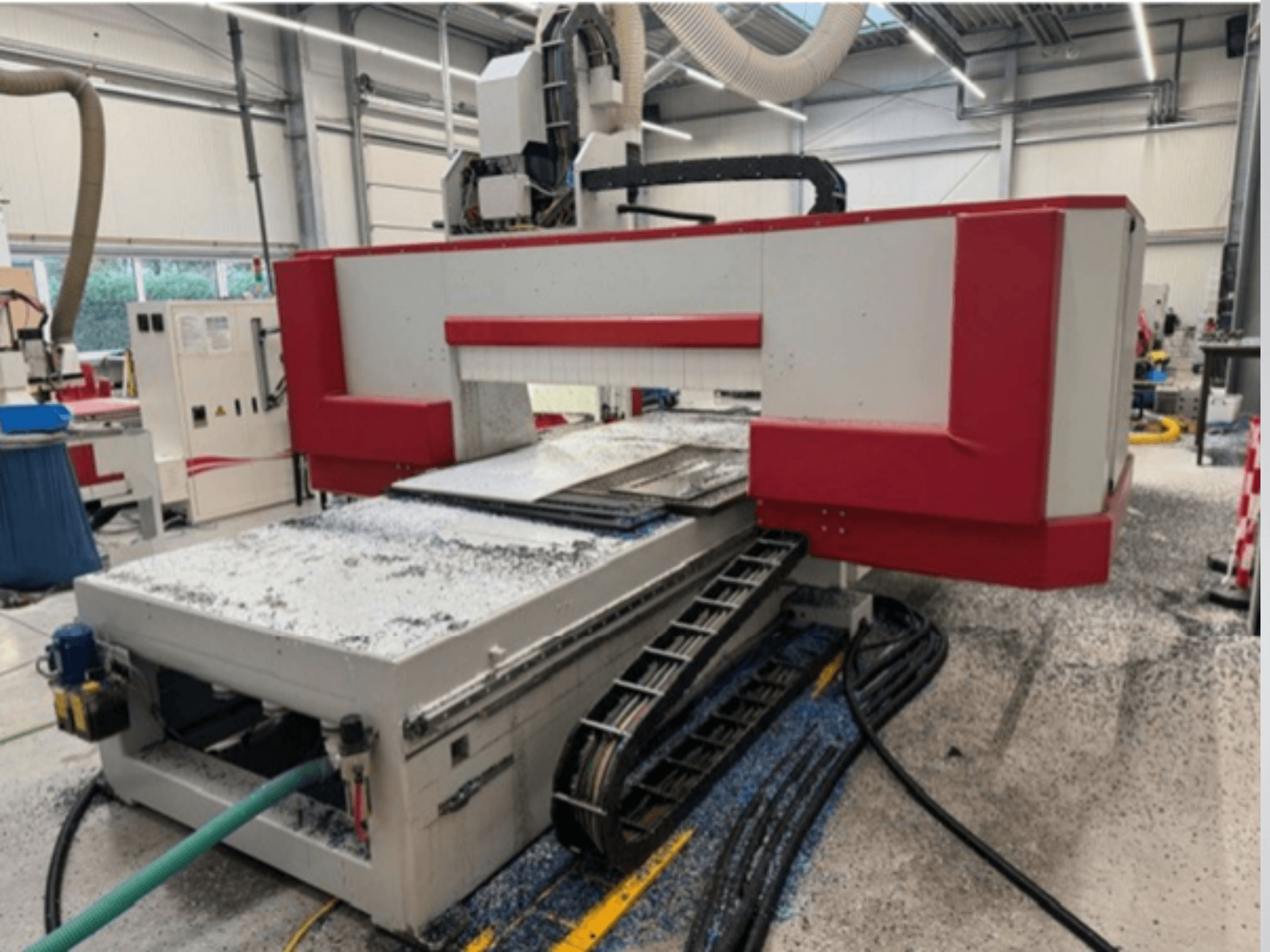 Punainen ja harmaa CNC-kone työpajassa, sivukuva, jossa näkyy materiaalipöytä, jäähdytysnesteletku ja sahanpuru lattialla.
