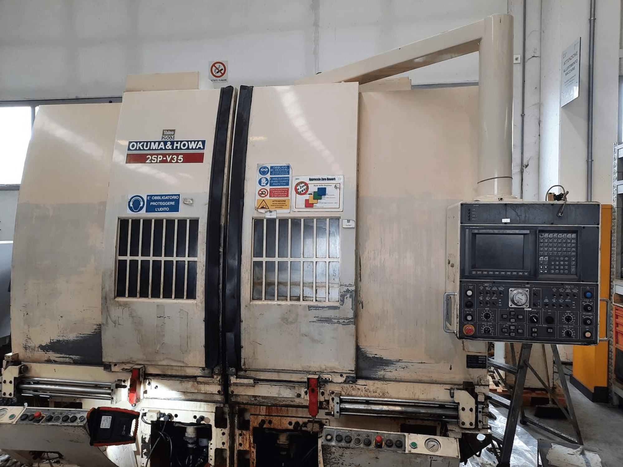 OKUMA & HOWA 2SP-V35 CNC-kone edestä katsottuna, jossa on ohjauspaneeli, jossa on erilaisia painikkeita ja turvatarroja.