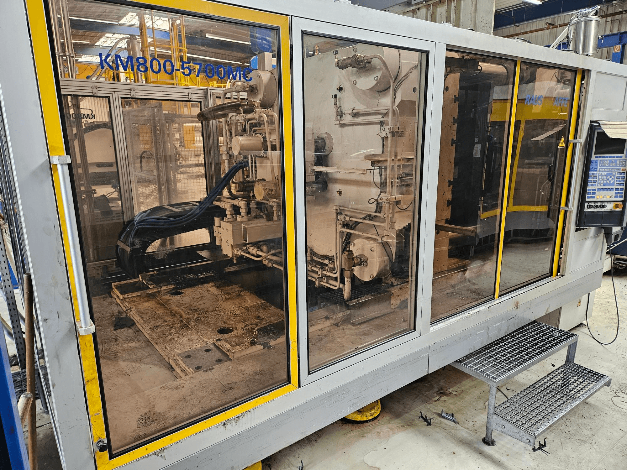 Krauss Maffei KM800-5700 MC -hydraulinen ruiskuvalukone, etunäkymä, jossa komponentit näkyvät lasipaneelien läpi.