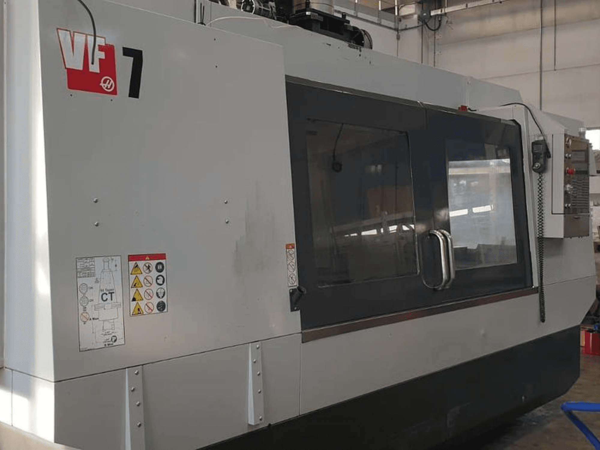 Haas VF-7 CNC-kone sivukuvassa, jossa näkyy metallinharmaa viimeistely, ohjauspaneeli ja turvatarrat.