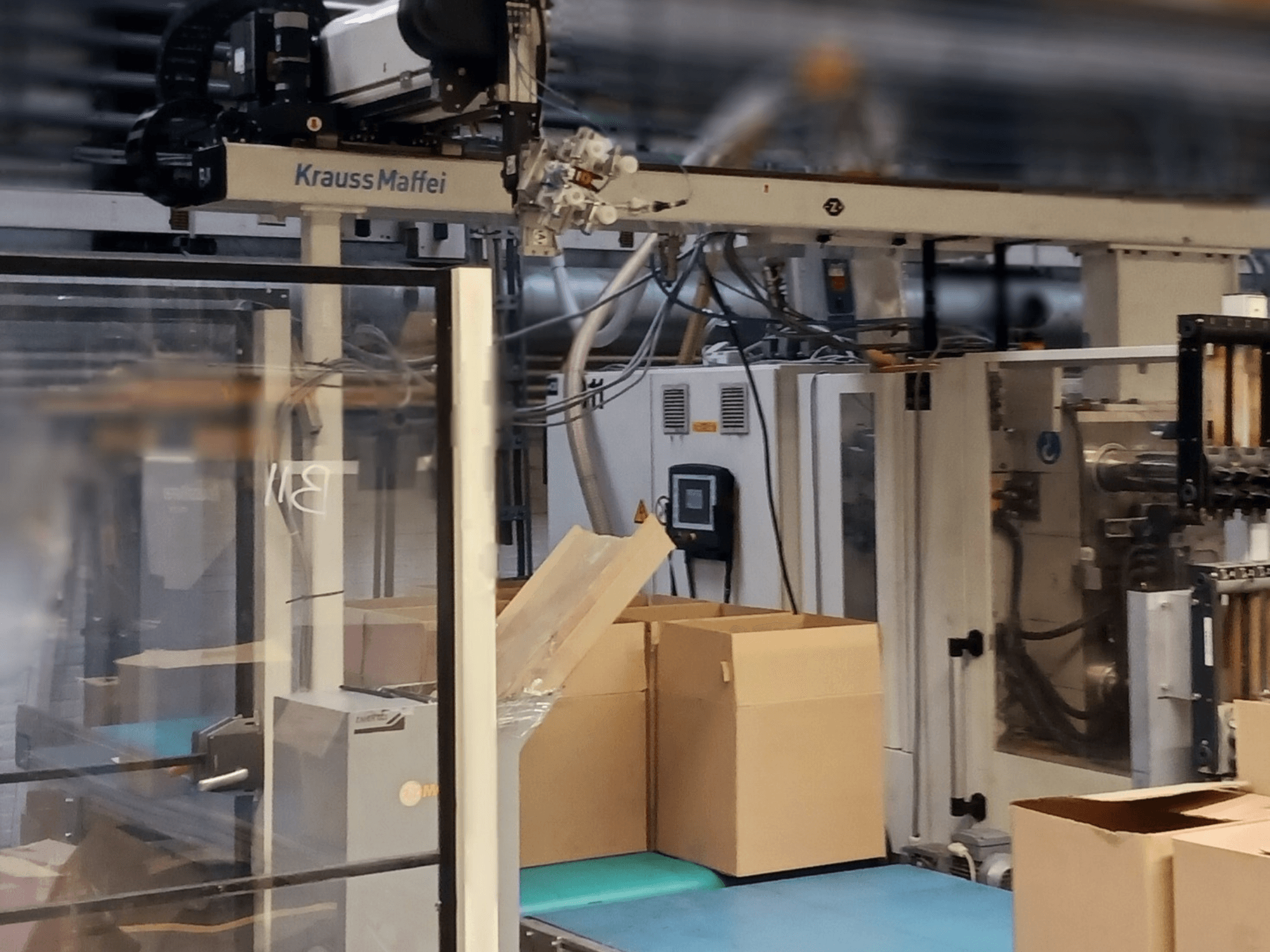 Krauss Maffei -automaatiojärjestelmä tehtaalla, etukuva, jossa näkyy laatikoita kuljettimella ja koneen osia toiminnassa.