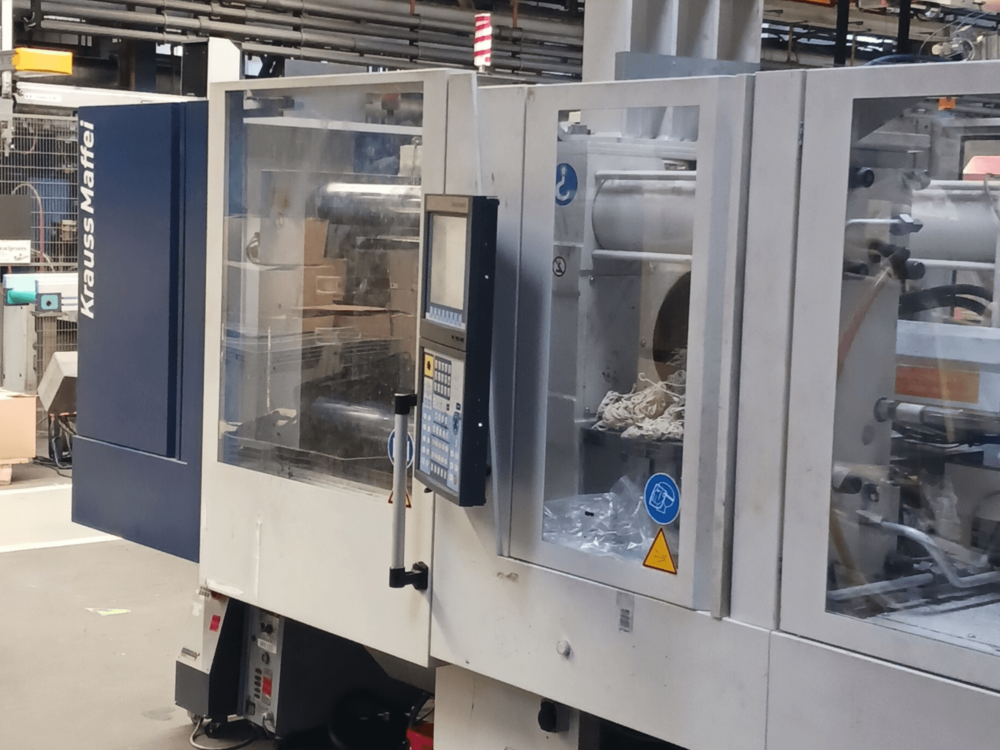 KraussMaffein ruiskuvalukone, sivukuva, jossa näkyy ohjauspaneeli, koneisto ja pakkausmateriaalit taustalla.