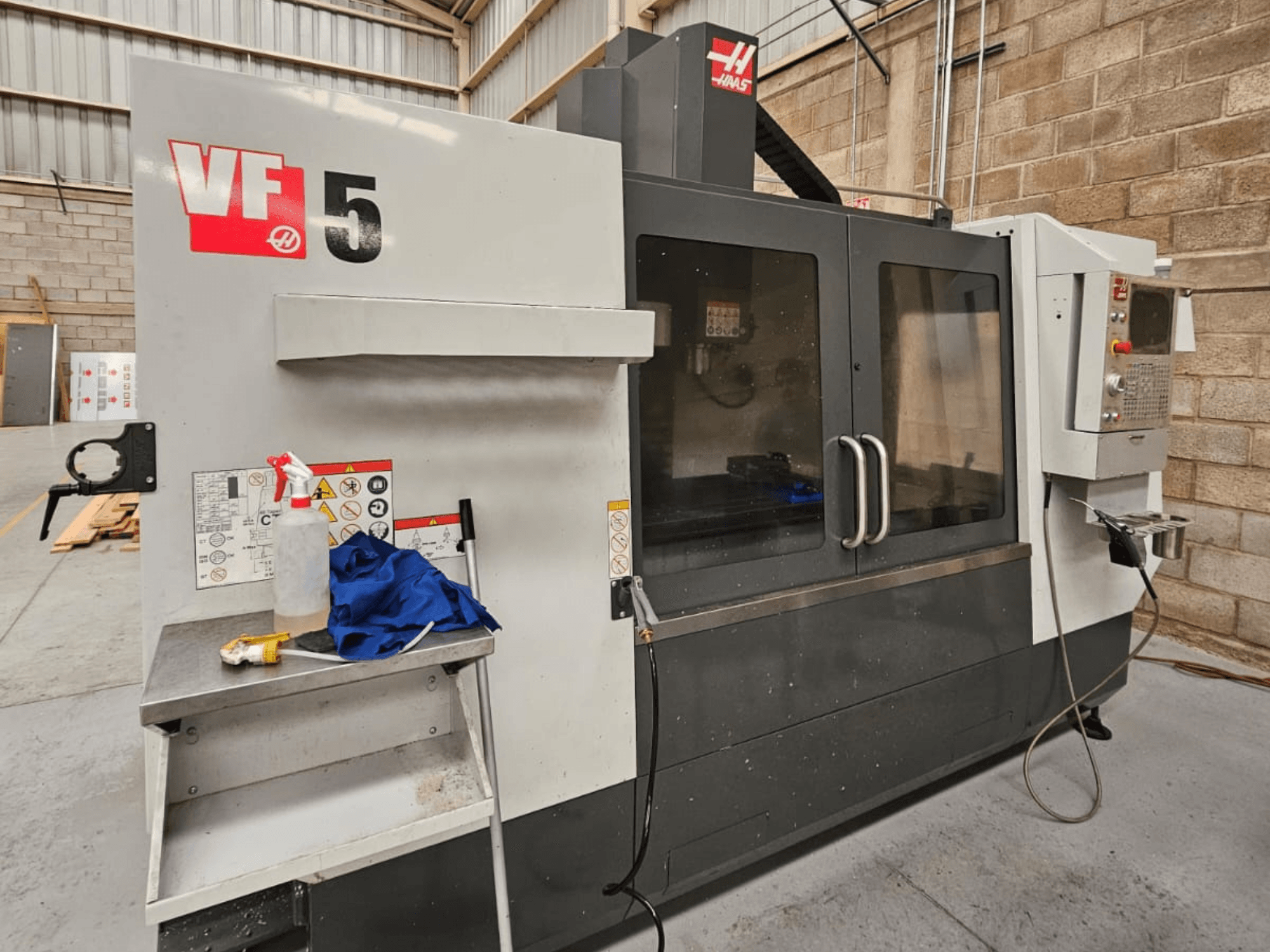 Haas VF-5 CNC-jyrsinkone etukuvassa, jossa on ohjauspaneeli, jäähdytysnestepullo, sininen liina ja turvaohjeet.
