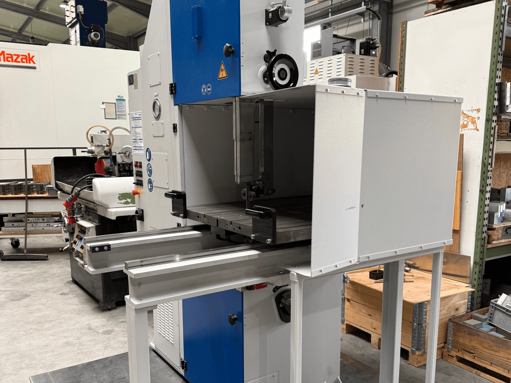 Mazak-teollisuuskone edestä katsottuna, jossa on sininen ohjauspaneeli, metallipöytä ja turvavaroitukset.