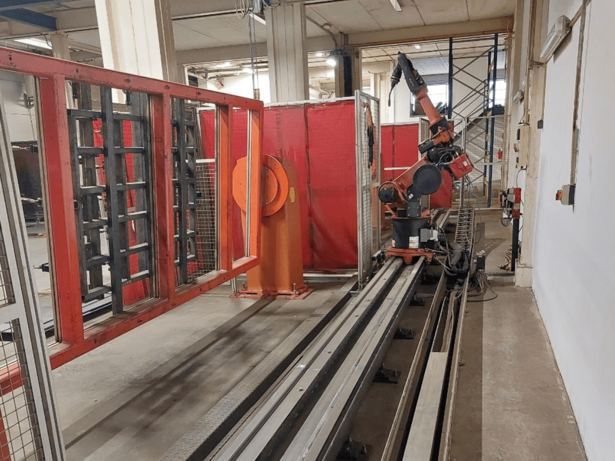 Oranssin ja mustan värinen teollisuusrobottikäsivarsi, joka on sijoitettu metallikiskoille ja jota ympäröivät punaiset suojakaiteet, etukuva.