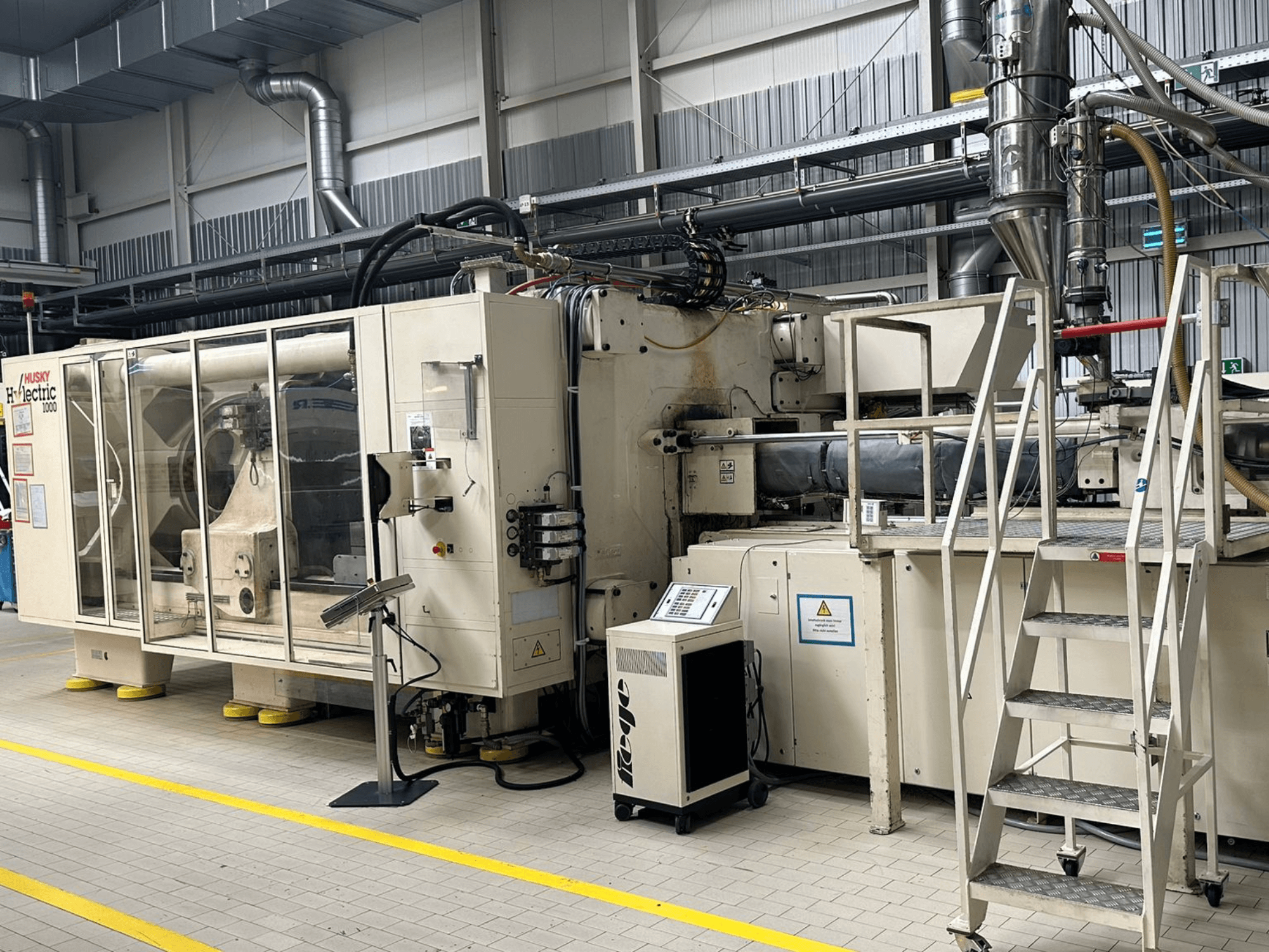 Husky Hylelectric H1000 RS95/80 -sähköisen ruiskuvalukoneen etunäkymä tehdasympäristössä; kuvassa näkyvät ohjauspaneeli ja portaat.