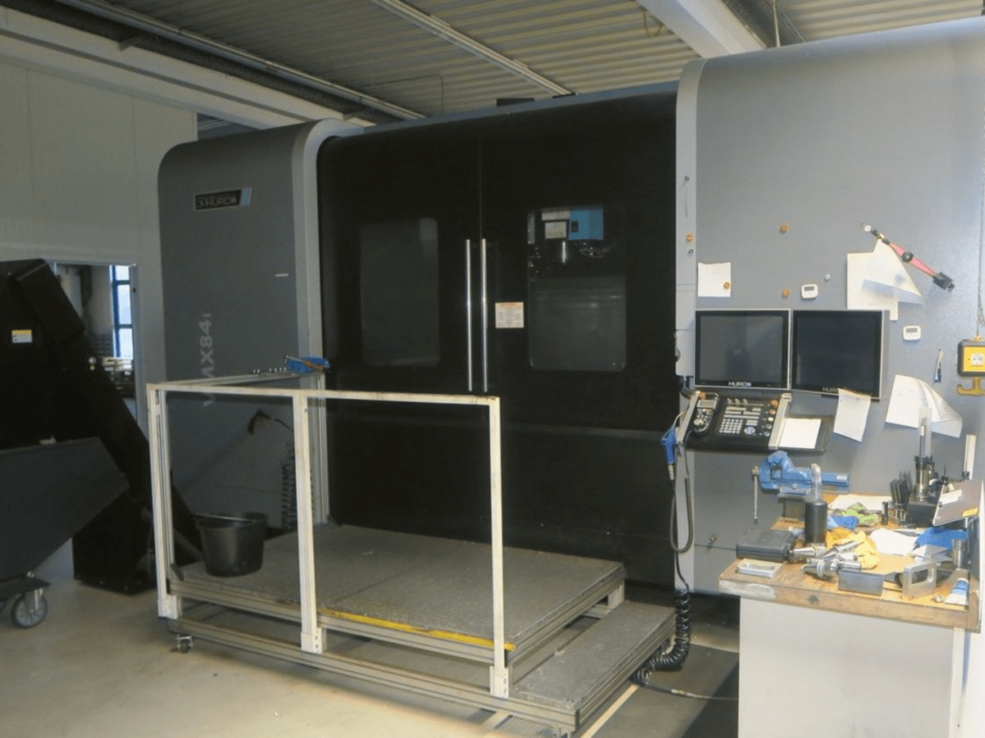 Hurco MX84 CNC-kone edestä katsottuna, jossa näkyy kaksoismonitorit, ohjauspaneeli ja työkalujen työtila, jossa on erilaisia työkaluja.