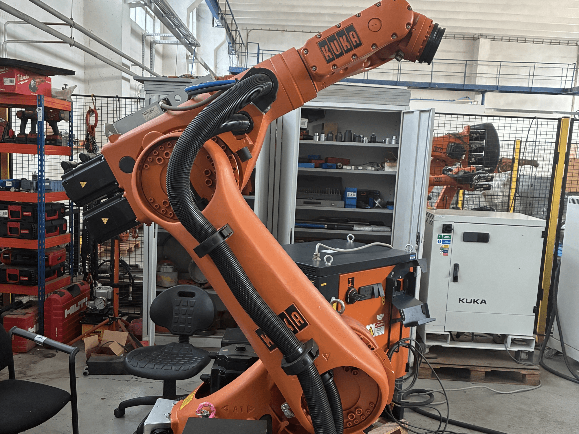 KUKA KR30-3 -robottivarsi teollisuusympäristössä, sivukuva, jossa näkyy kaapelit ja hallintalaitteet, taustalla työkaluja ja hyllyjä.