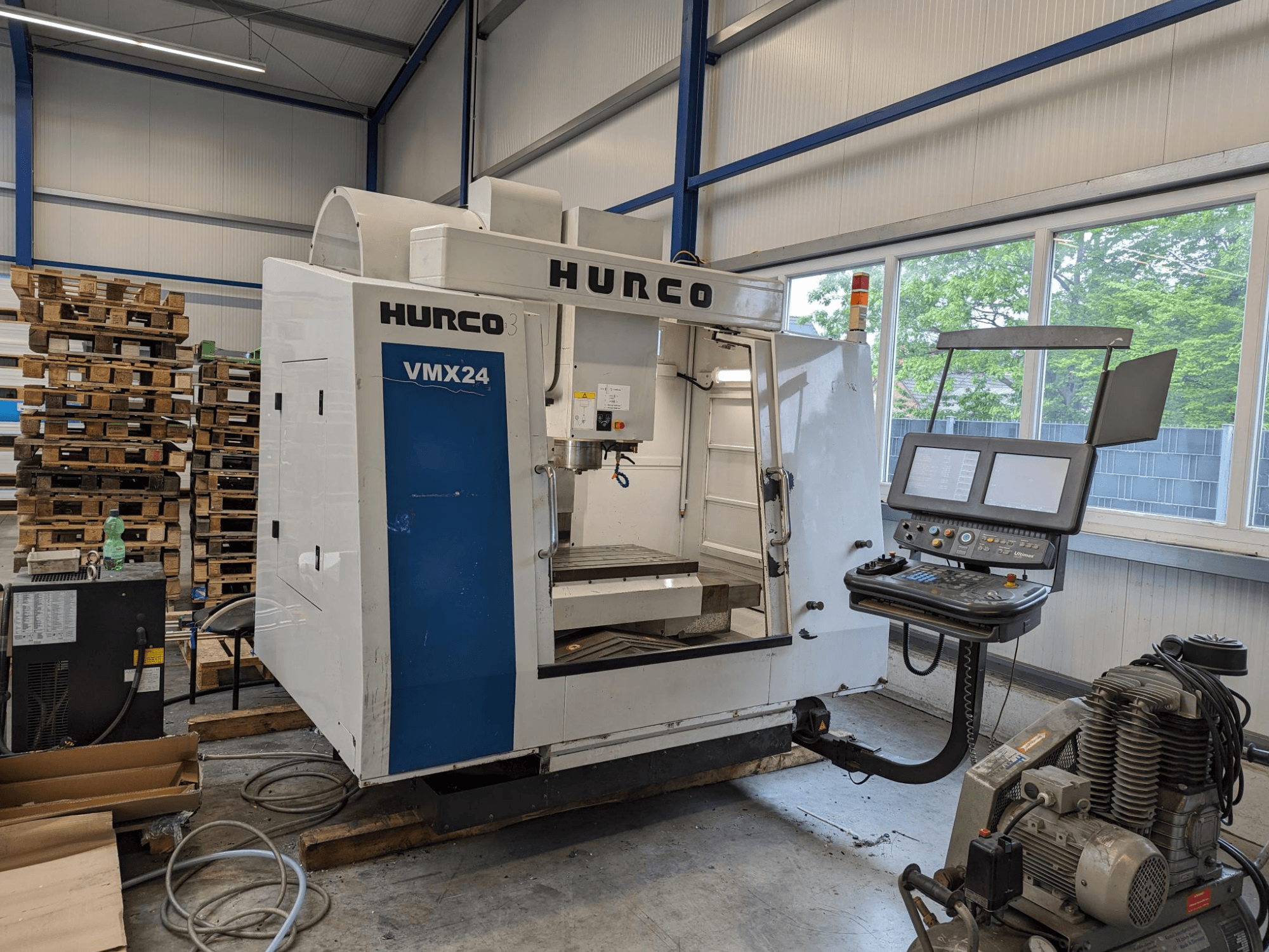 Hurco VMX24 -jyrsinkone, etukuva, jossa ohjauspaneeli ja työkalualue; taustalla pinottuja kuormalavoja; teollinen työtila-asetelma.