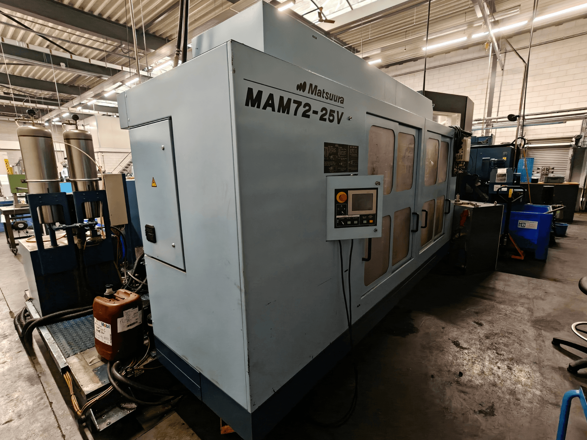 Matsuura MAM72-25V CNC-kone, sivukuva, jossa on ohjauspaneeli, kaksi jäähdytysnestesäiliötä ja työtila teollisuusympäristössä.