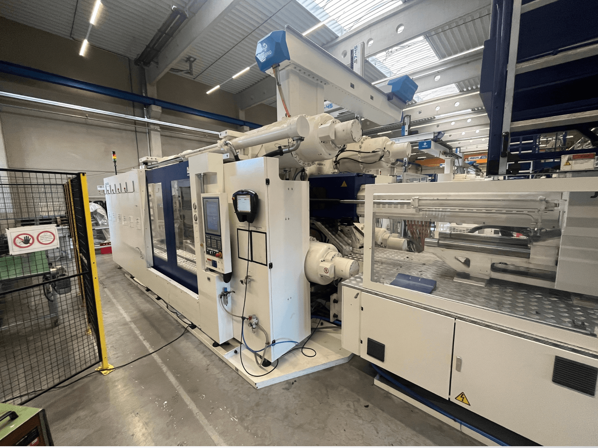 Valkoinen ja sininen ruiskuvalukone, etukuva, jossa on ohjauspaneeli, robottivarsi ja turvamerkit.