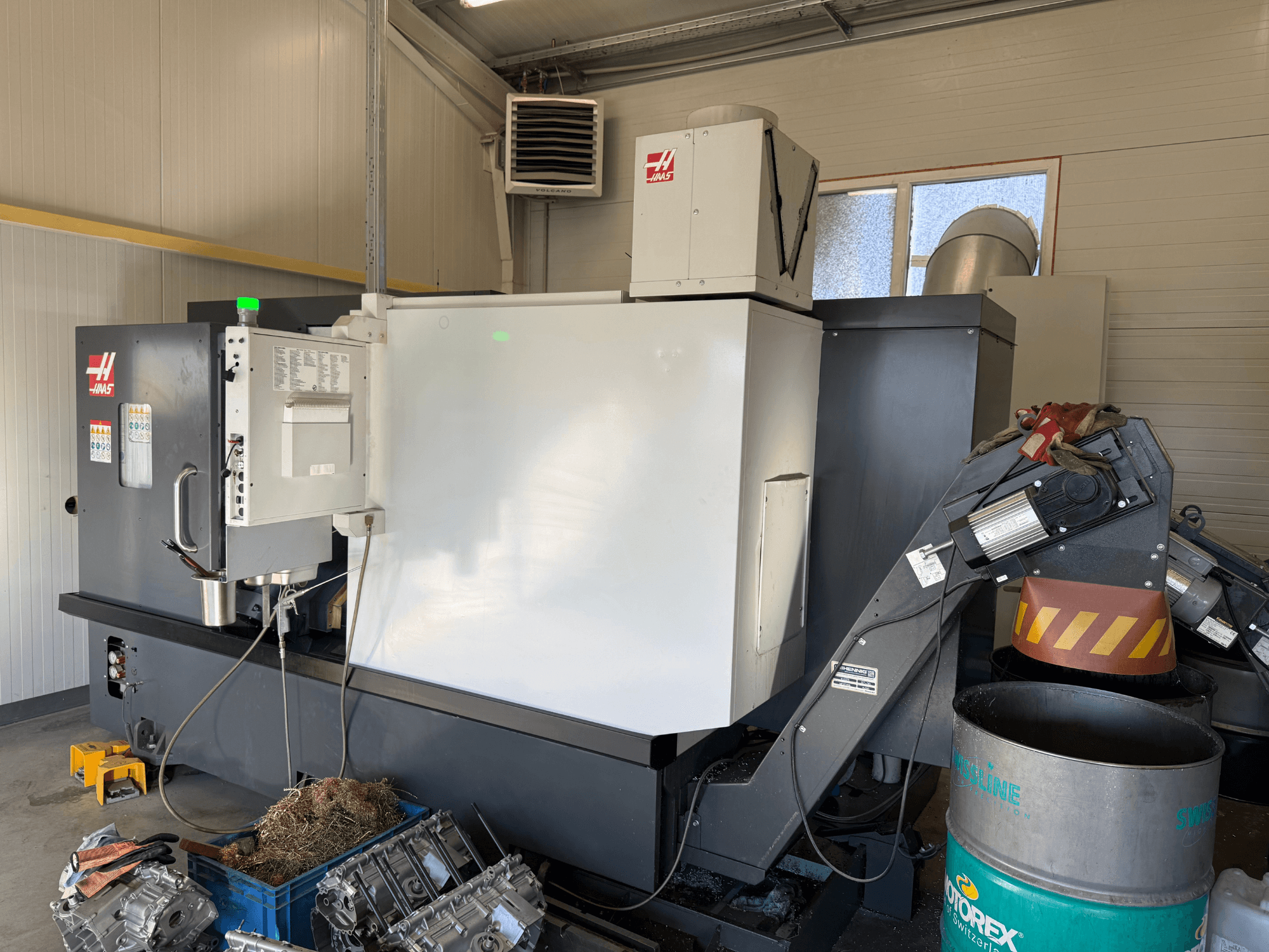 Haas CNC-kone ja ohjauspaneeli, sivukuva; näkyvät jäähdytysneste- ja öljylinjat; metalliromua sinisessä roskiksessa; teollisuusympäristö.