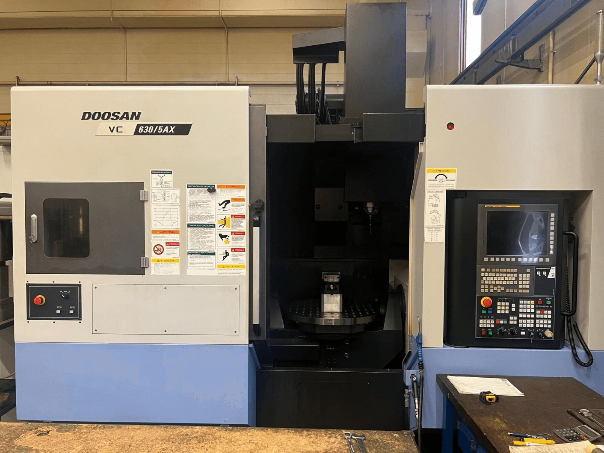 DOOSAN VC 630/5AX -pystykoneistuskeskuksen etunäkymä, jossa näkyvät ohjauspaneeli ja karan alue sekä turvallisuusmerkinnät.