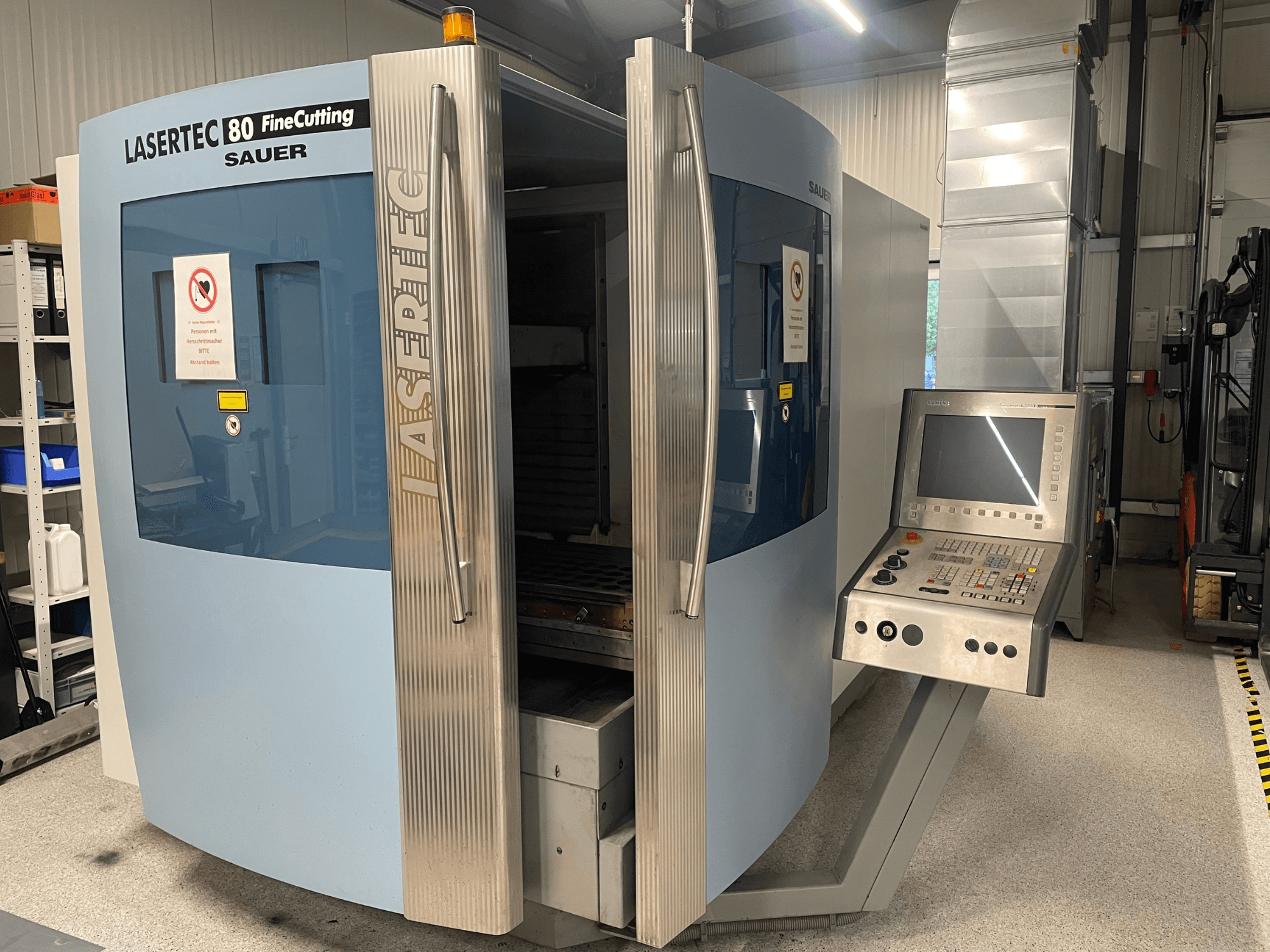 DMG Sauer Lasertec80 FineCutting -metallileikkauskoneen etunäkymä, jossa näkyy ohjauspaneeli, ovet auki ja turvallisuusmerkinnät.