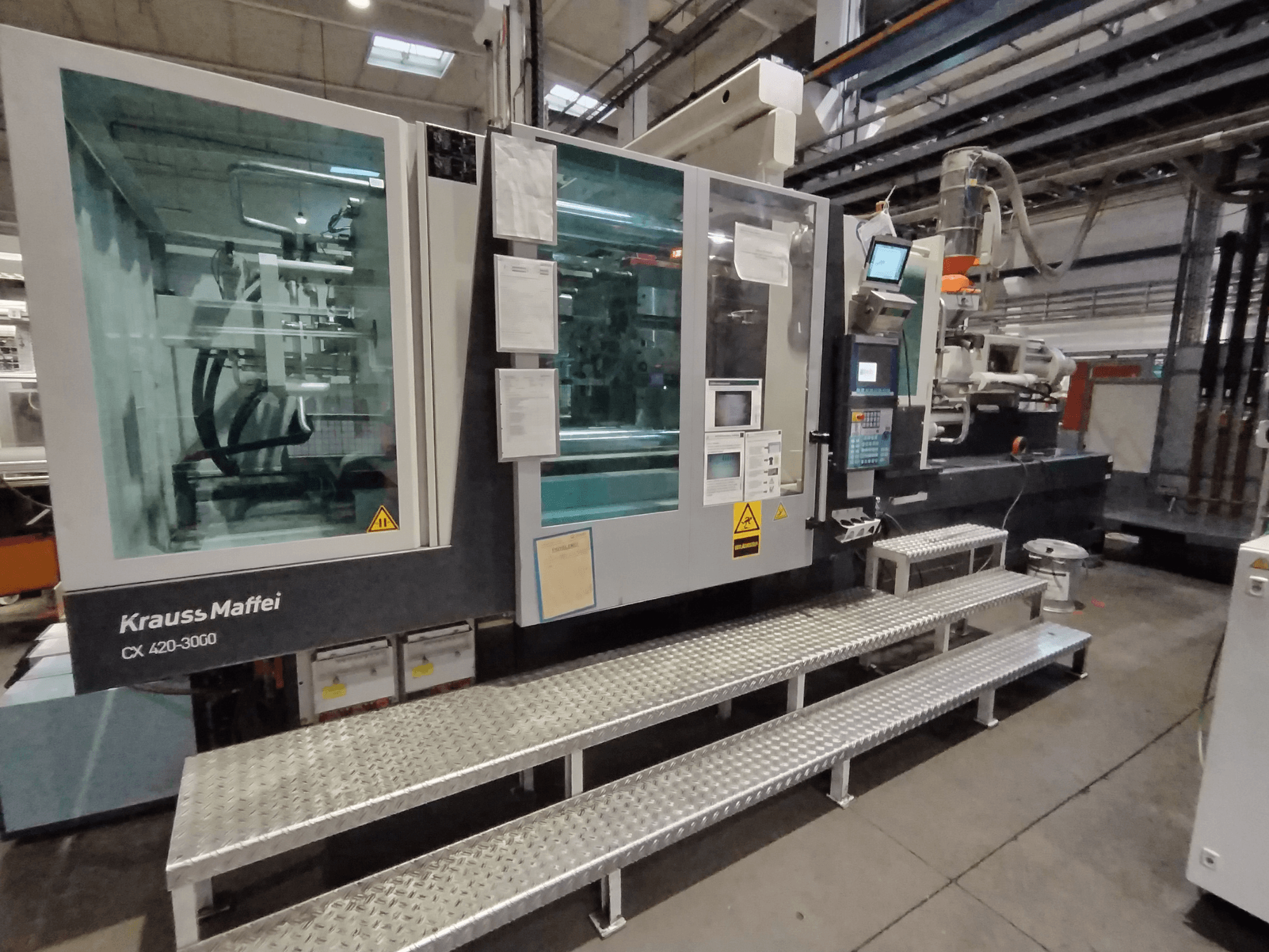 Krauss Maffei CX 420-3000 -ruiskuvalukone, sivukuva, ohjauspaneeli ja turvamerkinnät näkyvissä, metalliportaiden päällä.