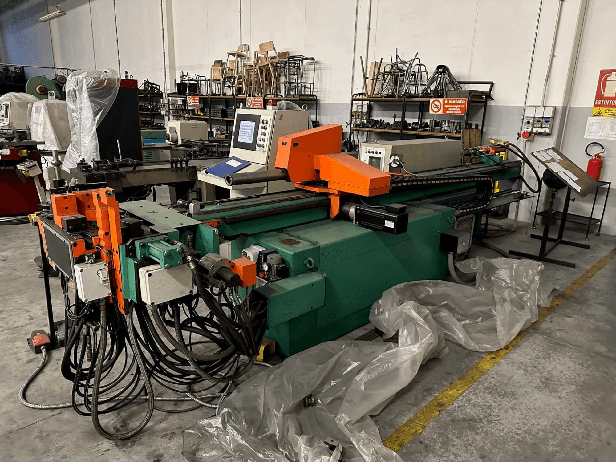 Teollisuuskone, jossa on oranssi ja vihreä runko, ohjauspaneeli ja lukuisia kaapeleita, edestä katsottuna työpajassa.