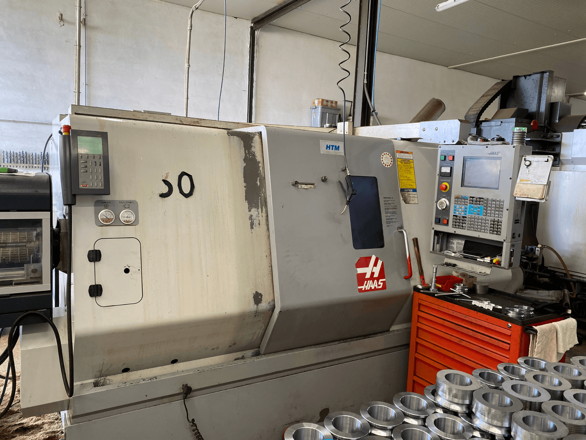 Haas CNC-kone, jossa on ohjauspaneeli, etukuva, jossa näkyvät käyttöpainikkeet, mittarit ja lähelle pinotut metalliosat.