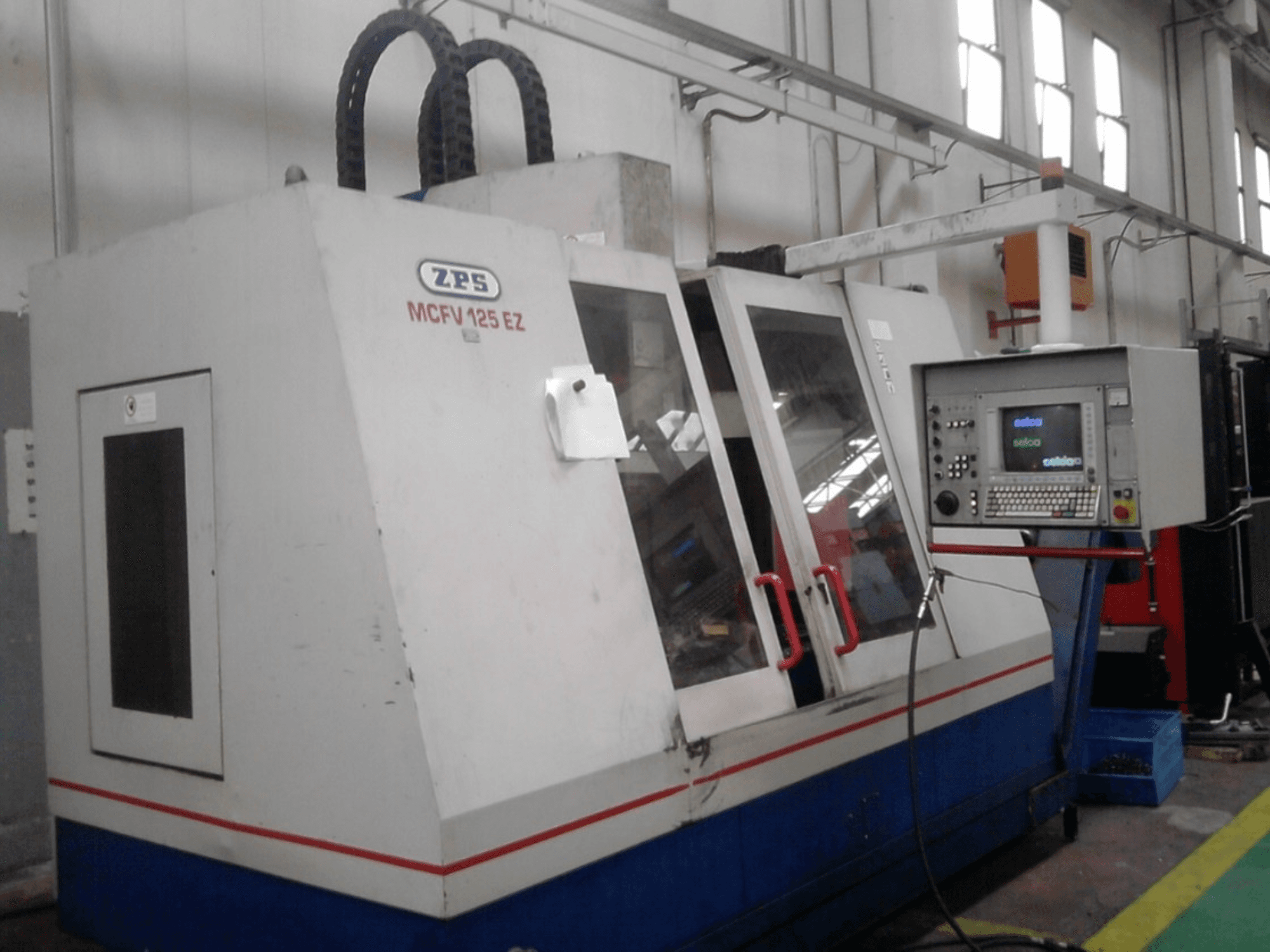 ZPS MCFV 125 EZ CNC-kone, etukuva, jossa ohjauspaneeli ja punaiset kahvat, työpajassa, jossa letkut ovat näkyvissä.