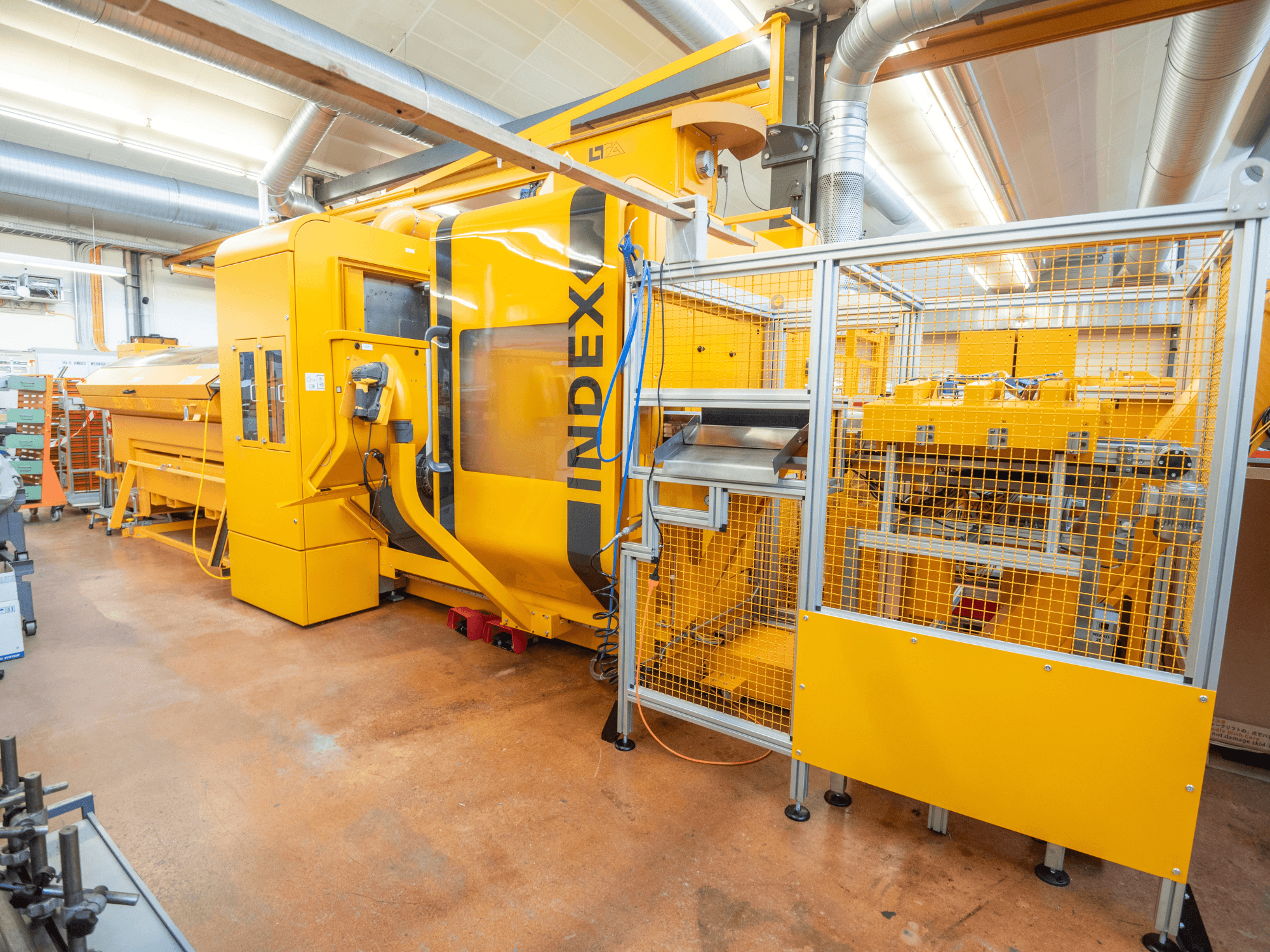 Keltainen INDEX-kone työpajassa, etukuva. Siinä on turvakotelo, ohjauspaneeli ja useita toiminnallisia osia.