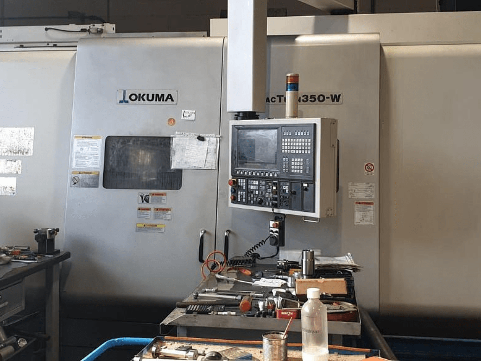 Okuma MAC TURN 350-W sorvauskeskus, etukuva, jossa ohjauspaneeli ja työtila täynnä työkaluja ja pullo.