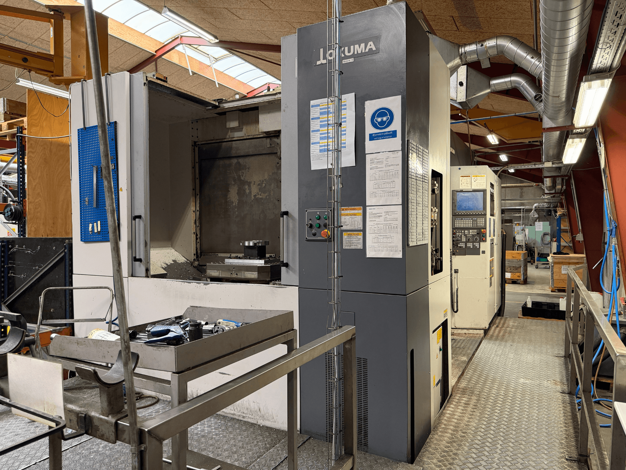 Okuma MA-600HB -vaakakoneistuskeskus, etunäkymä, jossa näkyvät metallipöydällä olevat työkalut ja koneen ohjauslaitteet.