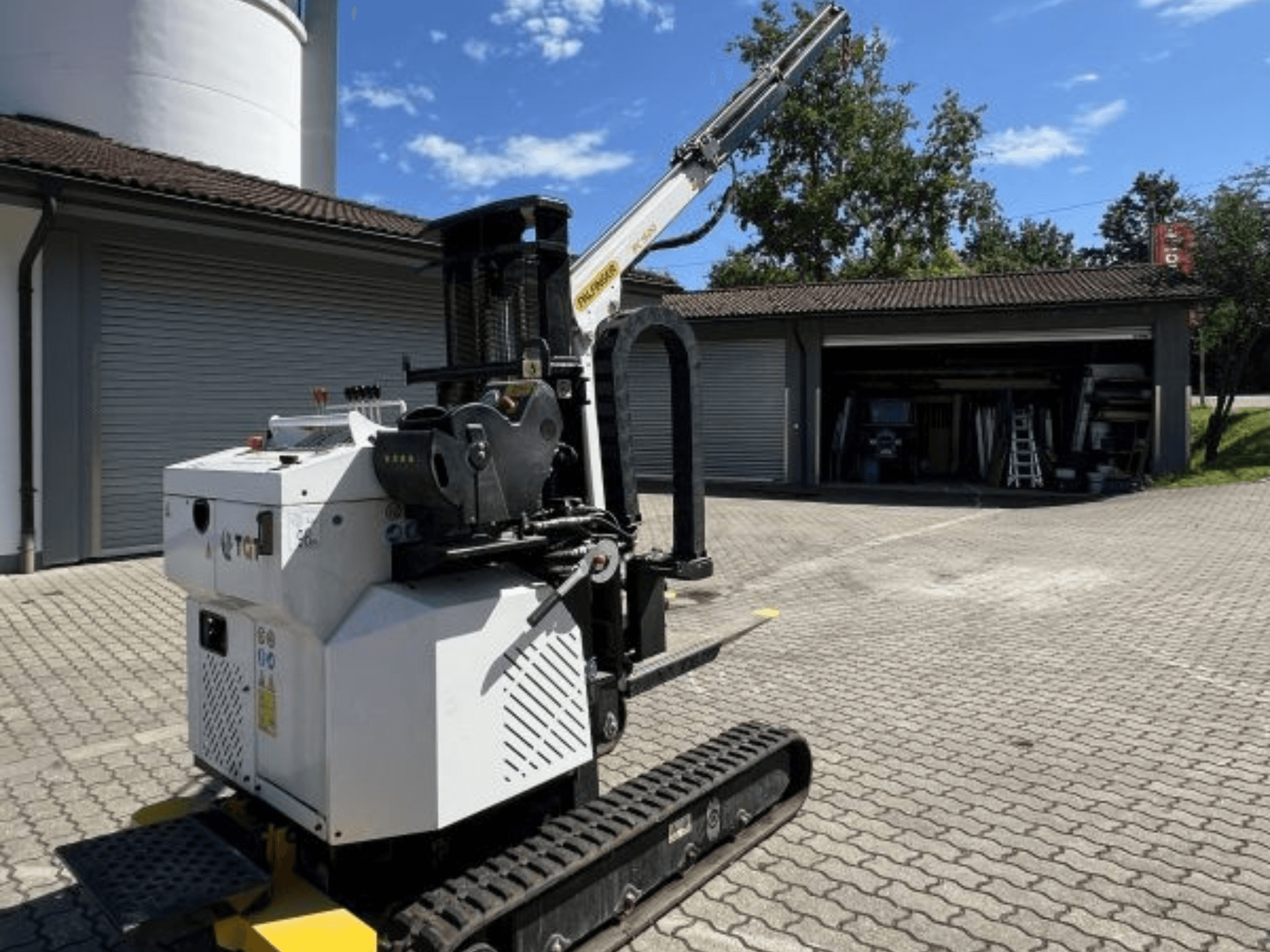 TGI-rakennusmininosturi, sivukuva, jossa näkyvät telaketjut ja asennettu puomi, pysäköity tiilipinnalle autotallin viereen.