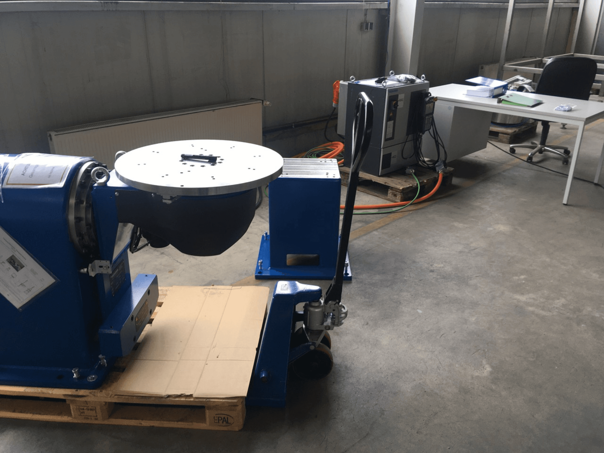 Sininen teollisuuslaite, jossa on pyöreä pohja ja ohjauspaneeli, edestä katsottuna puisella kuormalavalla toimistoympäristössä.