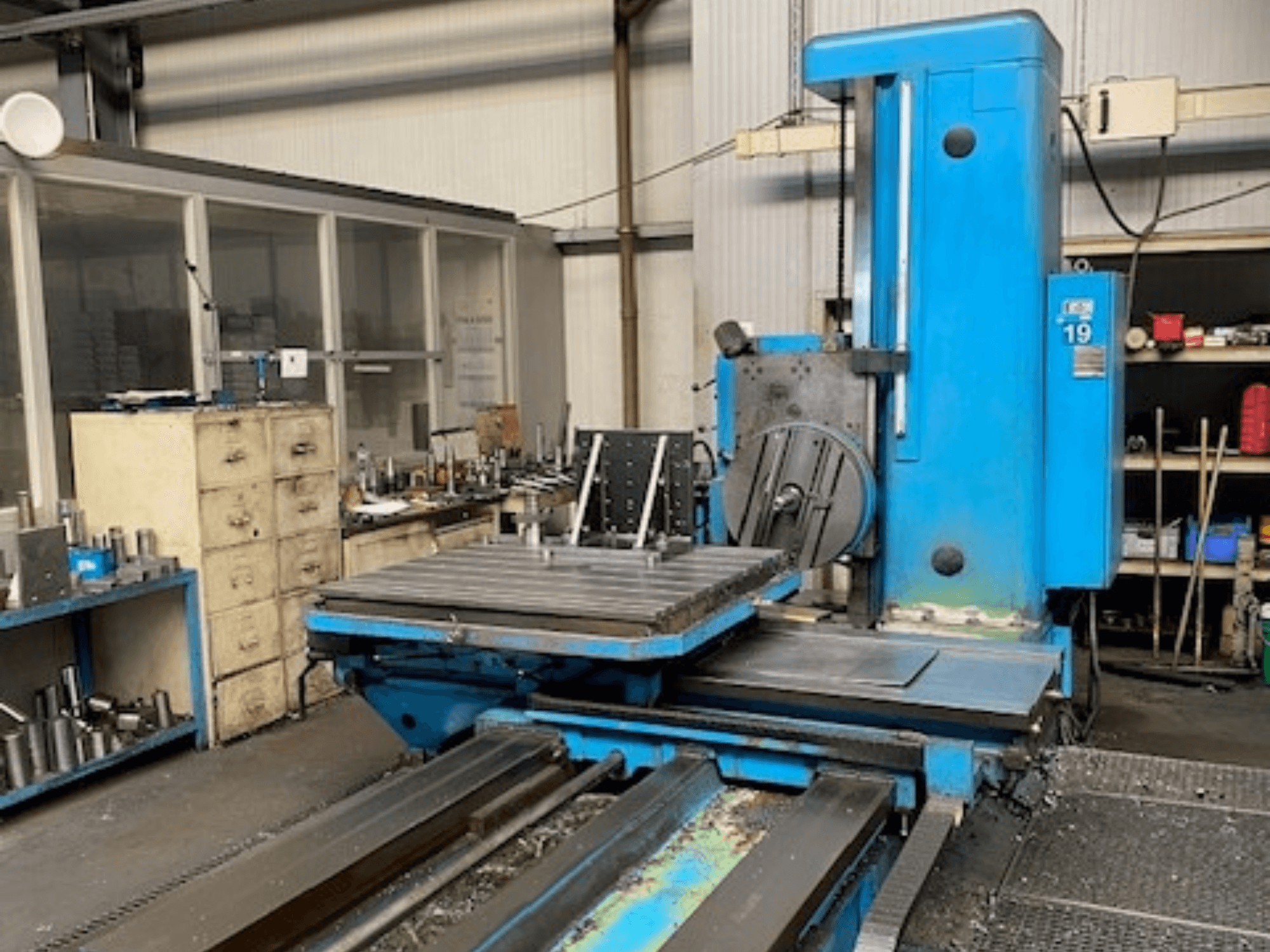 Sininen jyrsinkone etukuvassa, mallinumero 19, työpajan lattialla, taustalla työkaluja ja säilytyskaappi.