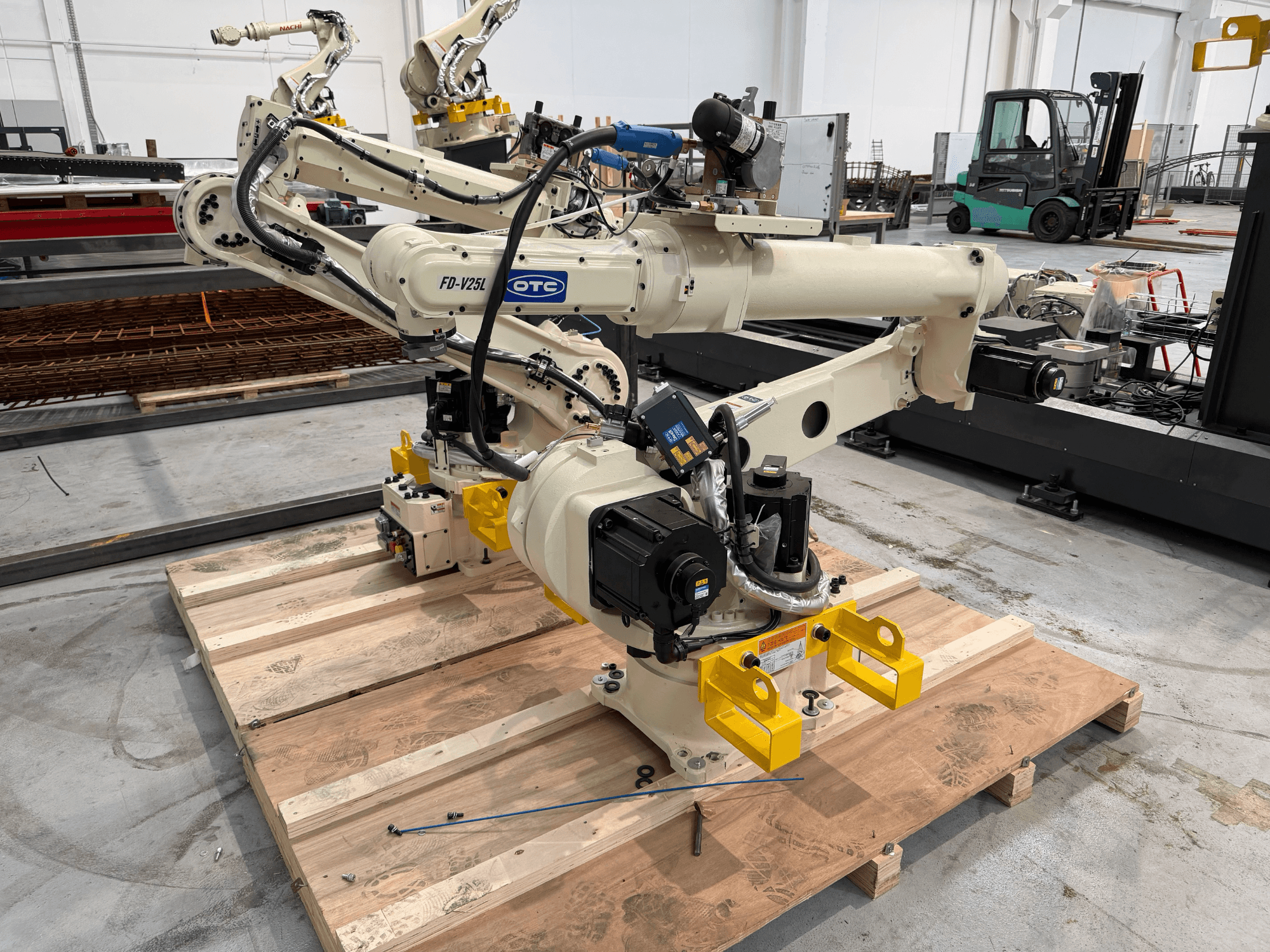 OTC Daihenin FD19-V25L-robottivarsi edestä katsottuna, keltaisilla tartuntakynsillä ja näkyvillä mekaanisilla osilla puupalettilla.