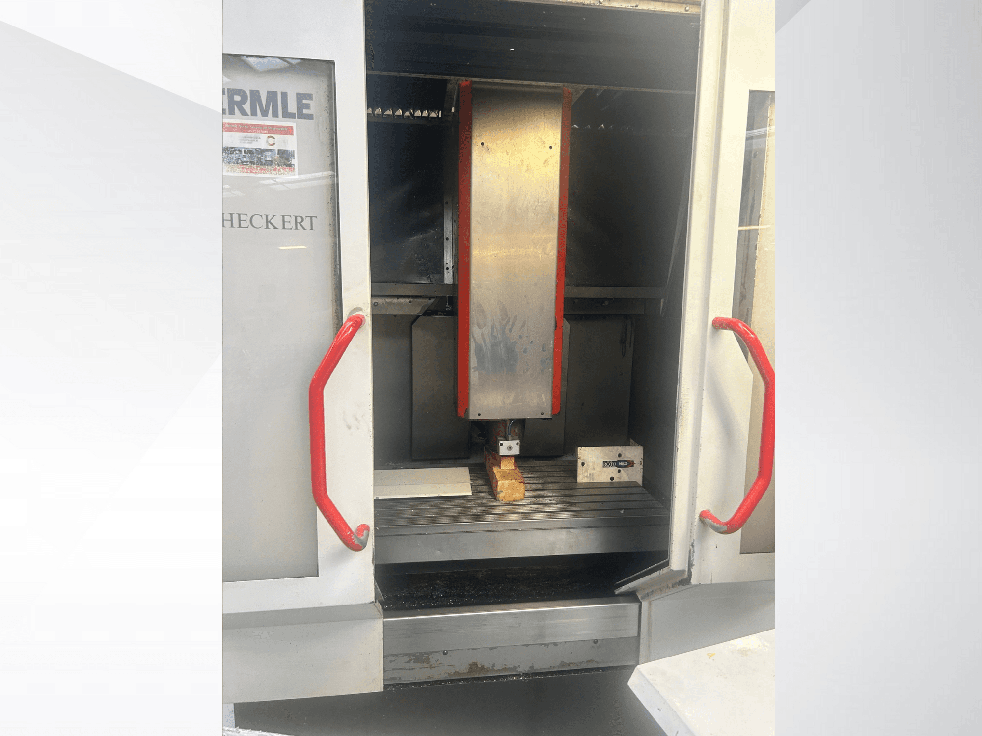 Hermle CNC-koneen etukuva, jossa on metallinen sisustus, punaiset kahvat ja työkappale pöydällä.