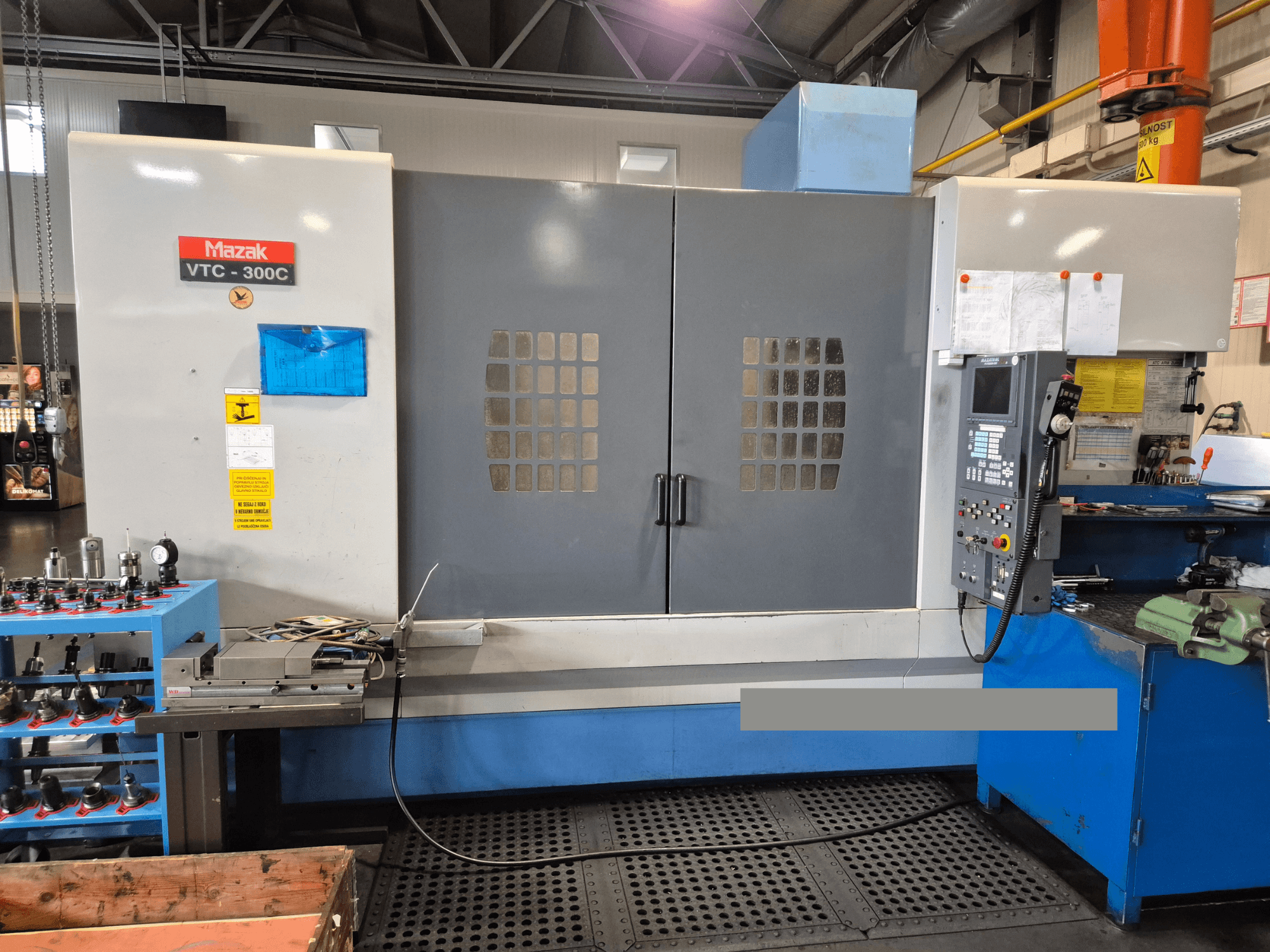 Mazak VTC-300C -kone tehtaalla, etukuva, jossa näkyy ohjauspaneeli, harmaat ovet ja ikkunaruutu sekä lähellä olevat laitteet.