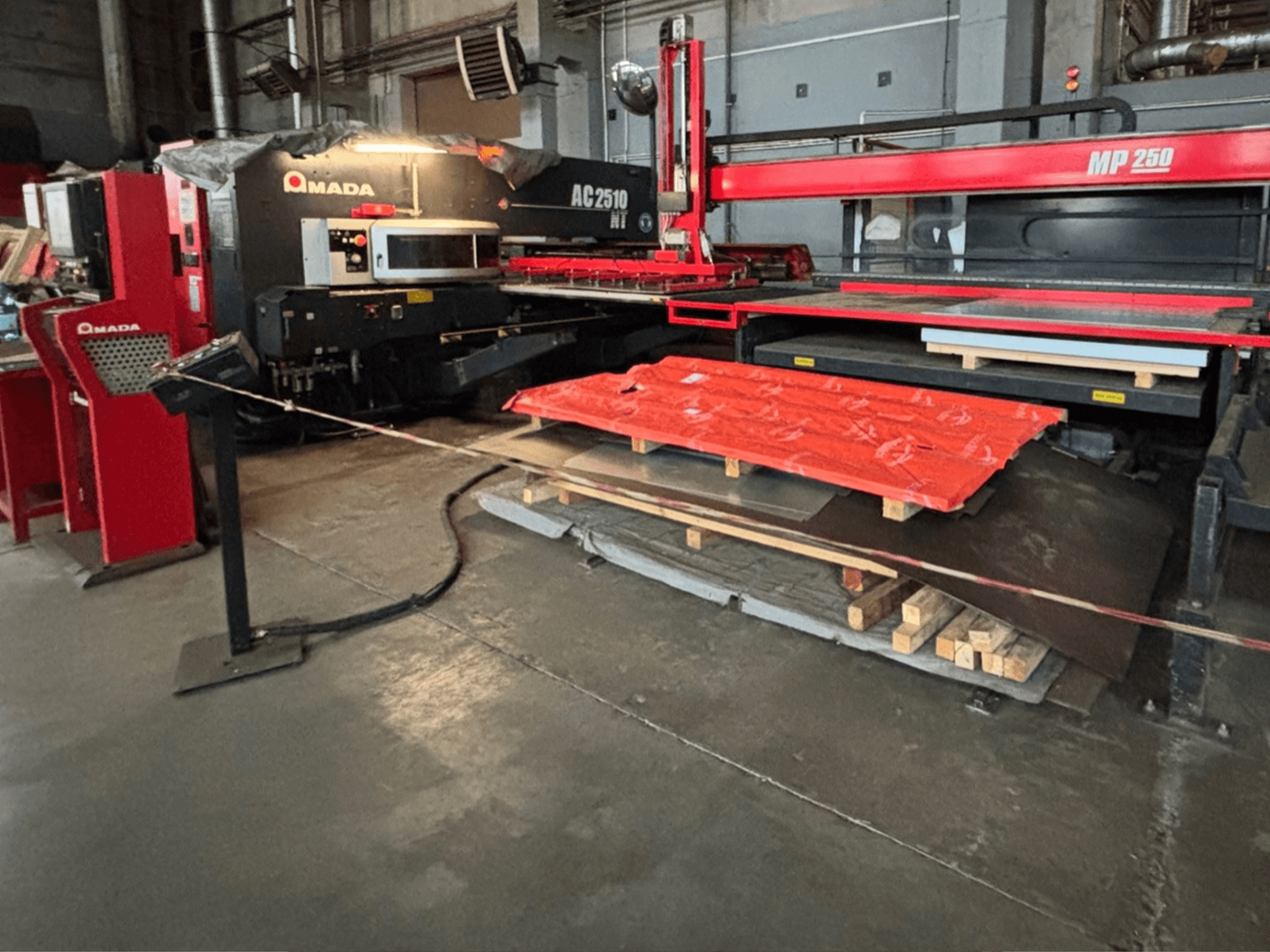 Amada AC2510 NT -laserleikkuri tehdasympäristössä, etukuva, raskas koneisto, lattialla metallilevyjä ja kuormalavoja.