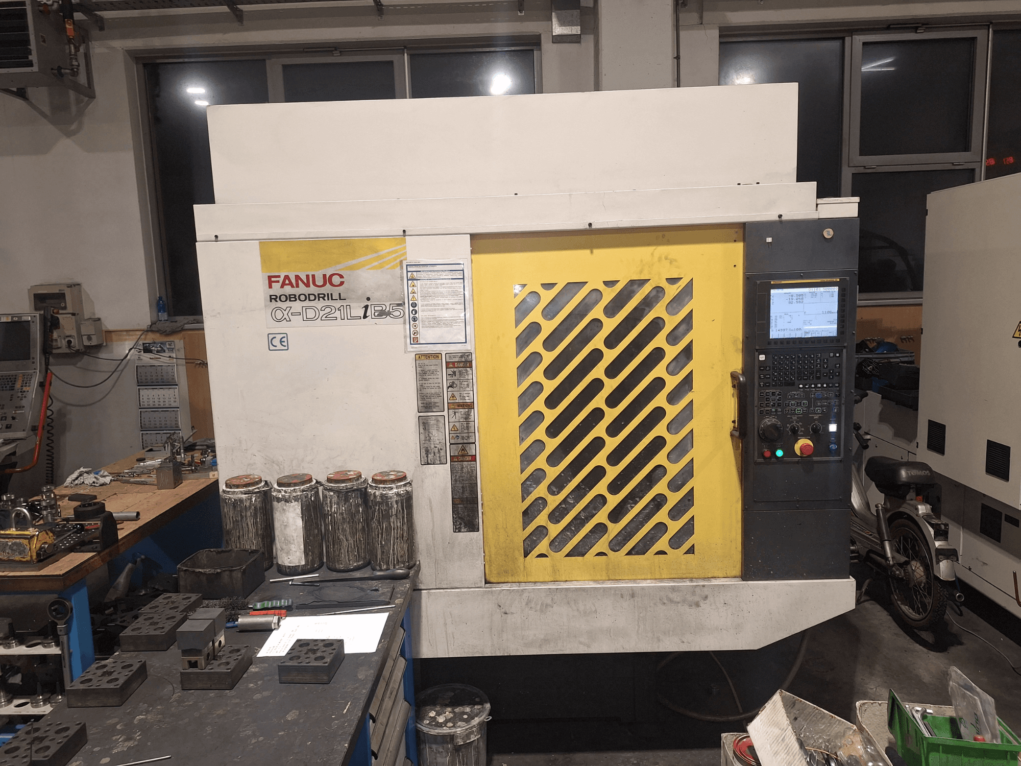 FANUC Robodrill α-D21LiB5:n etukuva, jossa näkyy ohjauspaneeli, keltainen säleikkö ja läheiset työkalut työpöydällä.