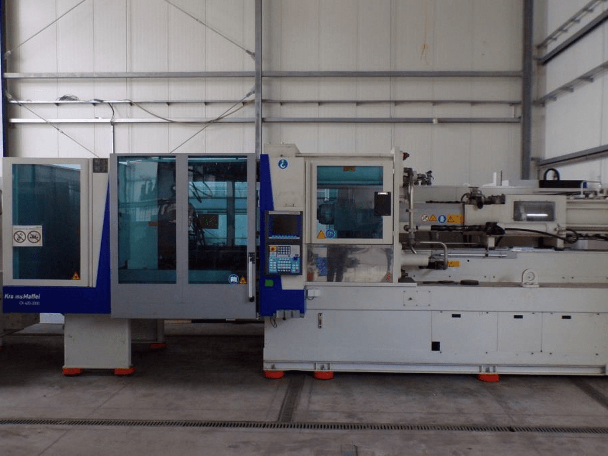KraussMaffei ruiskuvalukone, sivukuva, jossa on ohjauspaneeli ja turvamerkinantolaitteet, jotka näkyvät tehdasympäristössä.