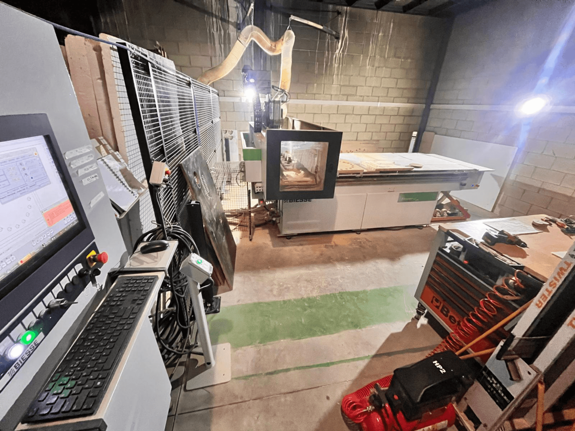 Biesse CNC-kone työpajassa, sivukuva, jossa näkyy ohjauspaneeli, pölynpoisto, työkalut ja erilaisia materiaaleja pöydällä.