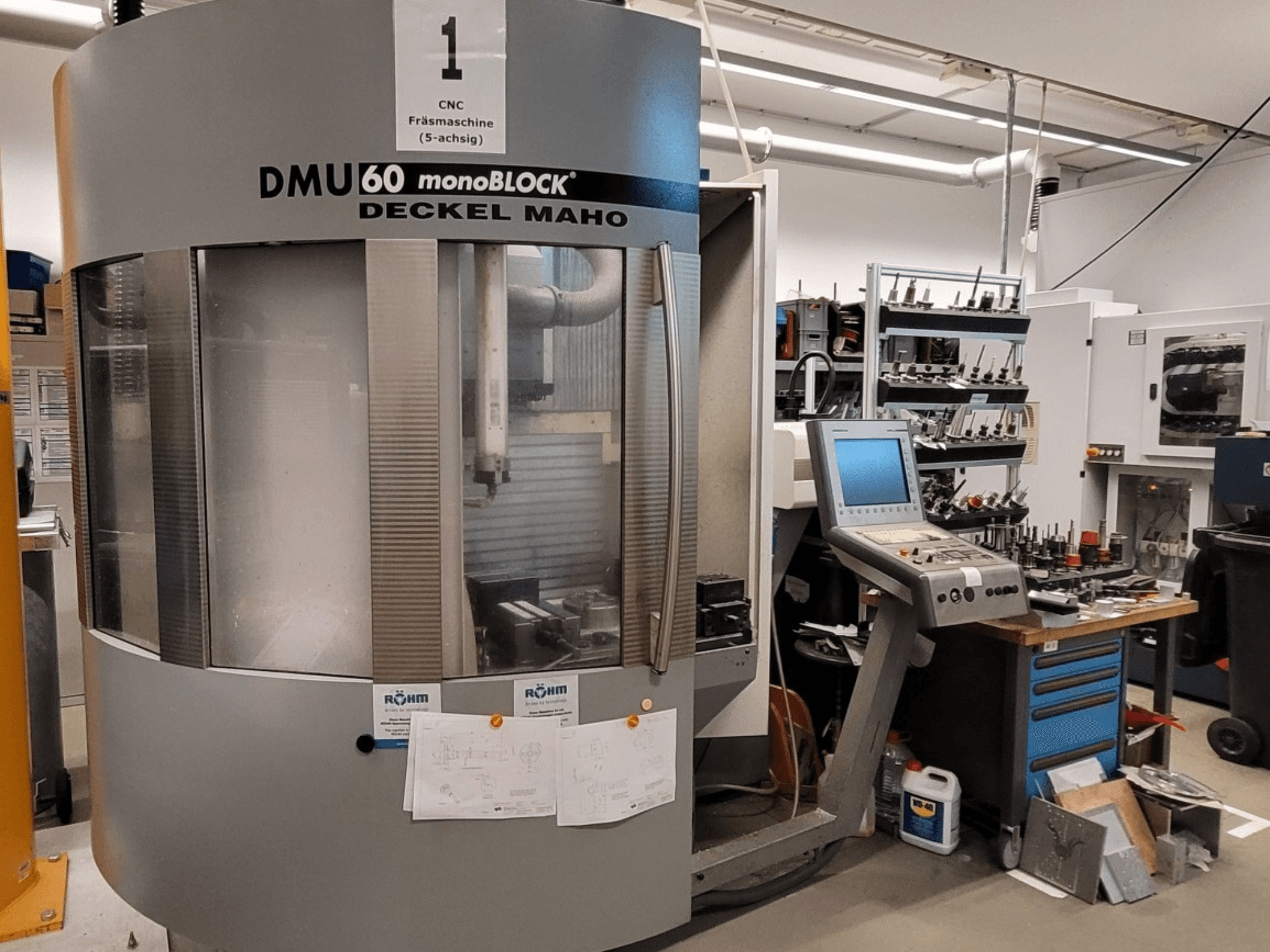 CNC-jyrsinkone DECKEL MAHO DMU 60 monoBLOCK etukuvassa, jonka vieressä on ohjauspaneeli ja työkalujen säilytys.