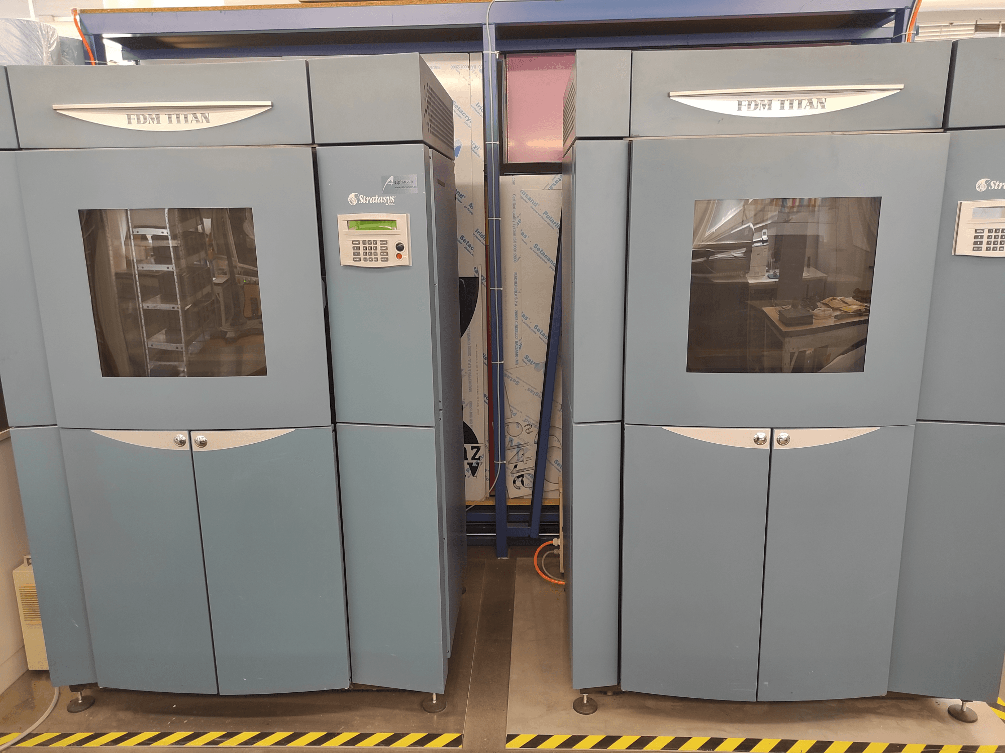 Stratasysin FDM TitanTI 3D-tulostin, etunäkymä, jossa näkyvät kaksoisovet ja ohjauspaneeli, sijoitettuna teollisuusympäristöön.