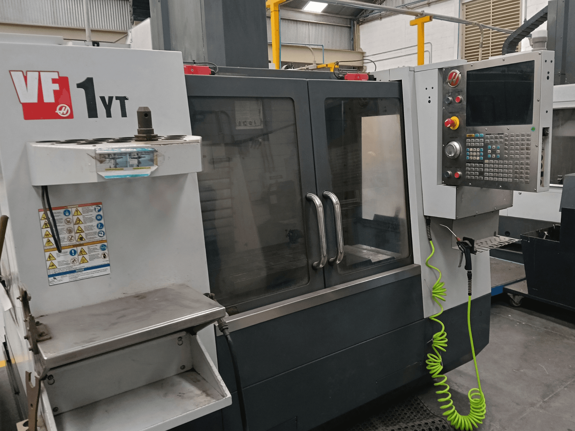 HAAS VF-1YT-2 -pystykoneistuskeskus, etunäkymä, jossa näkyvät ohjauspaneeli, työkalunpidin, turvallisuusmerkinnät ja koneistusalue.