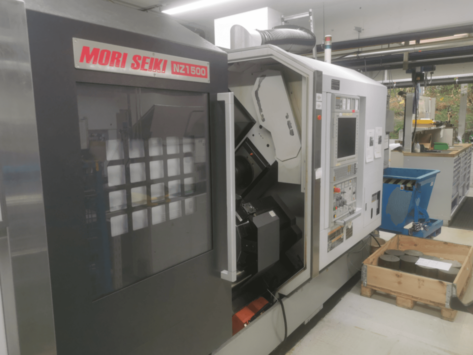 MORI SEIKI NZ1500 CNC-kone pienessä kulmassa, jossa näkyy ohjauspaneeli ja työtila, jossa on metallisia työkappaleita puisella kuormalavalla.