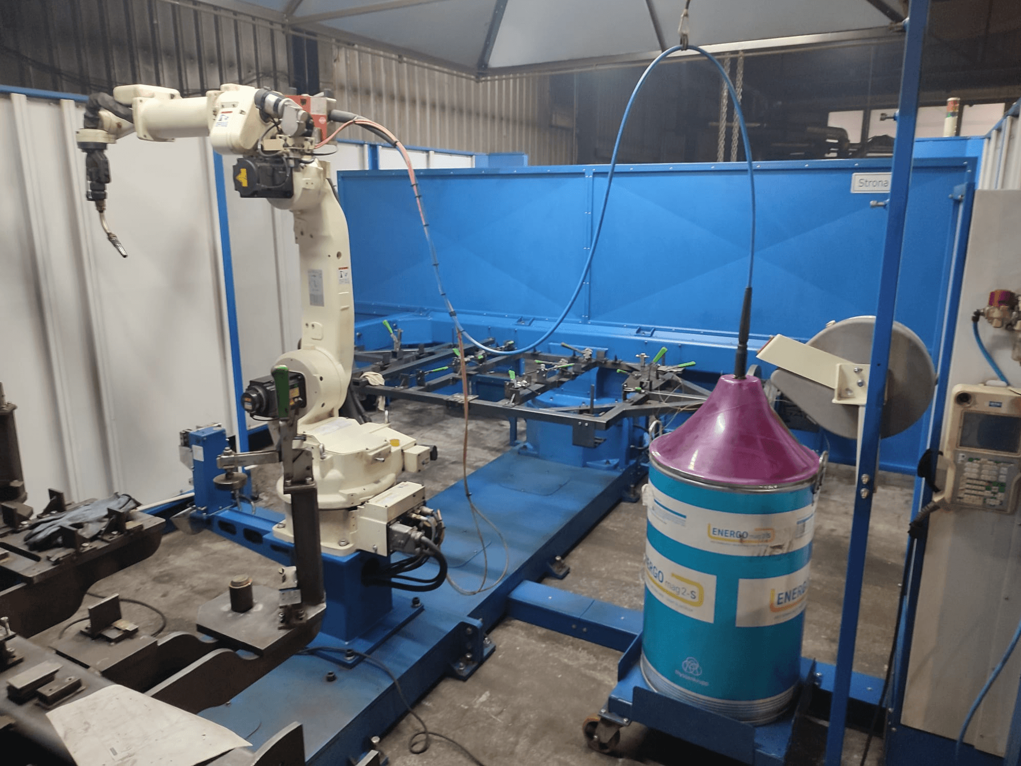 Teollisuusrobottivarsi, jossa on valkoinen ja sininen pohja, asennettuna työpajassa, jossa on sininen työtaso ja metallisia komponentteja.