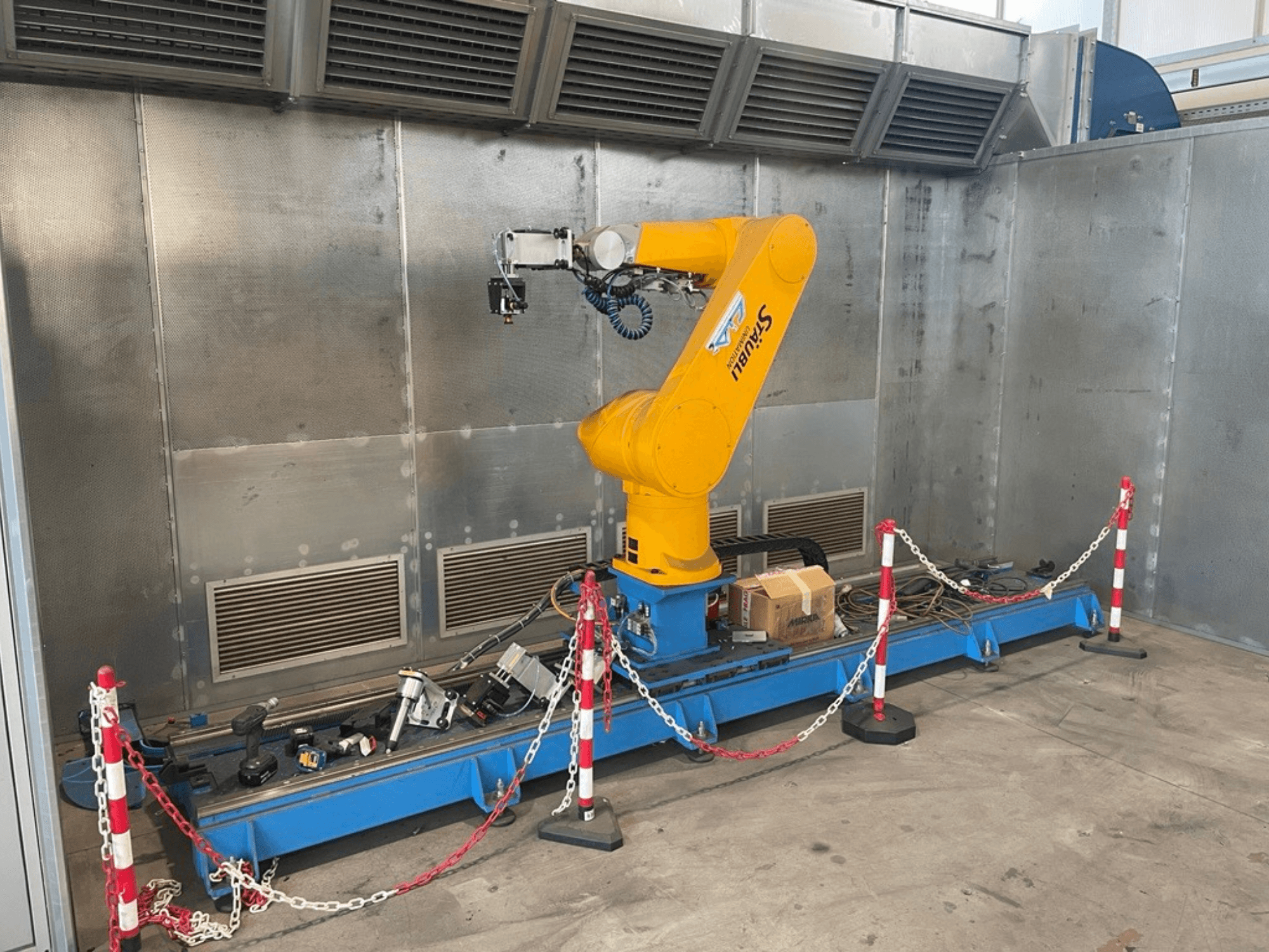 STAUBLI CS8C-RX160 -robottikäsivarsi työpajassa, sivukuva, kaapelit ja erilaiset työkalut sinisellä alustalla, jota ympäröivät suojakaiteet.
