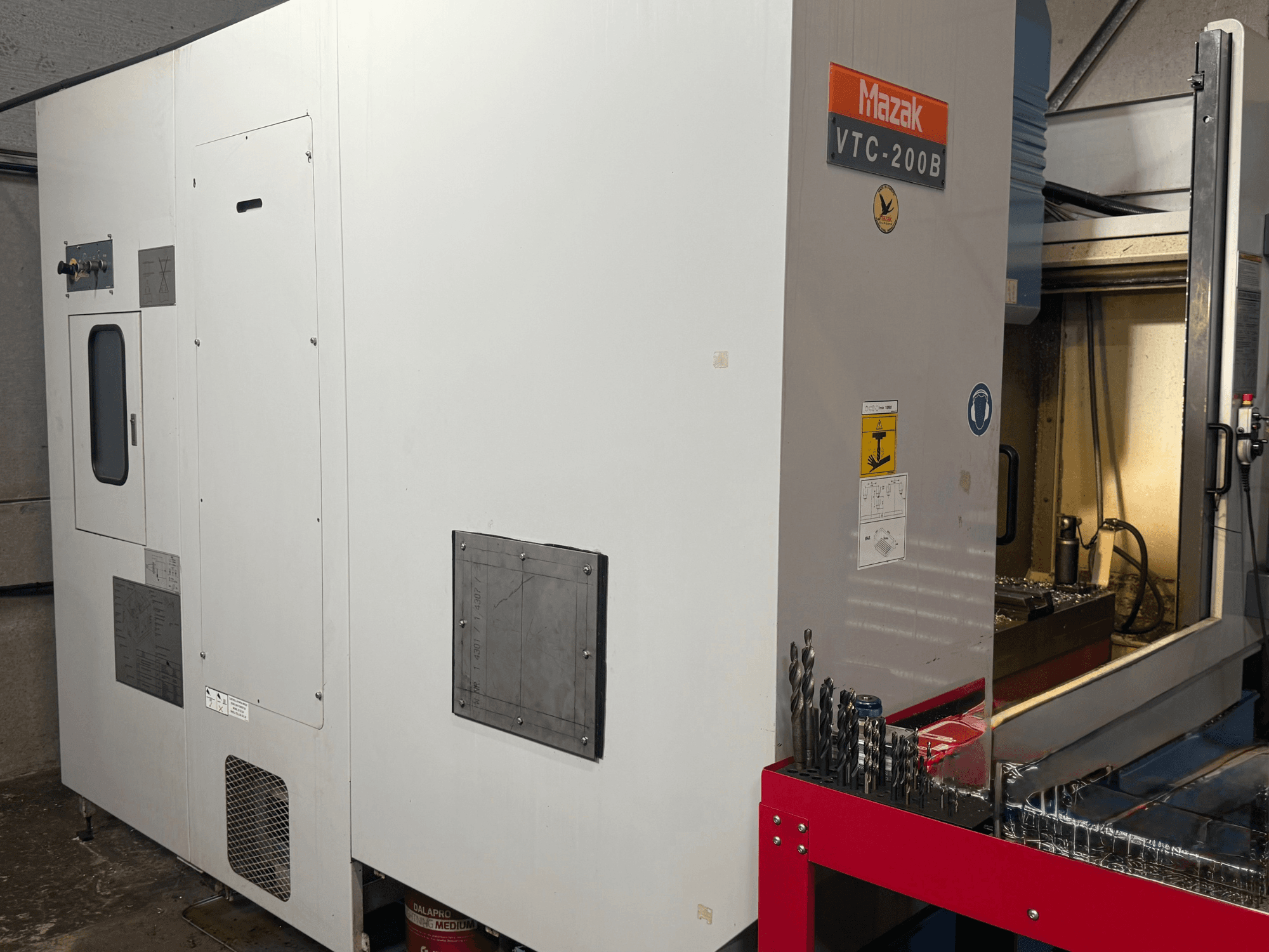 Mazak VTC-200B -pystykoneistuskeskus sivulta katsottuna; kuvassa näkyvät merkityt hallintalaitteet ja poranterät punaisella säilytystelineellä.