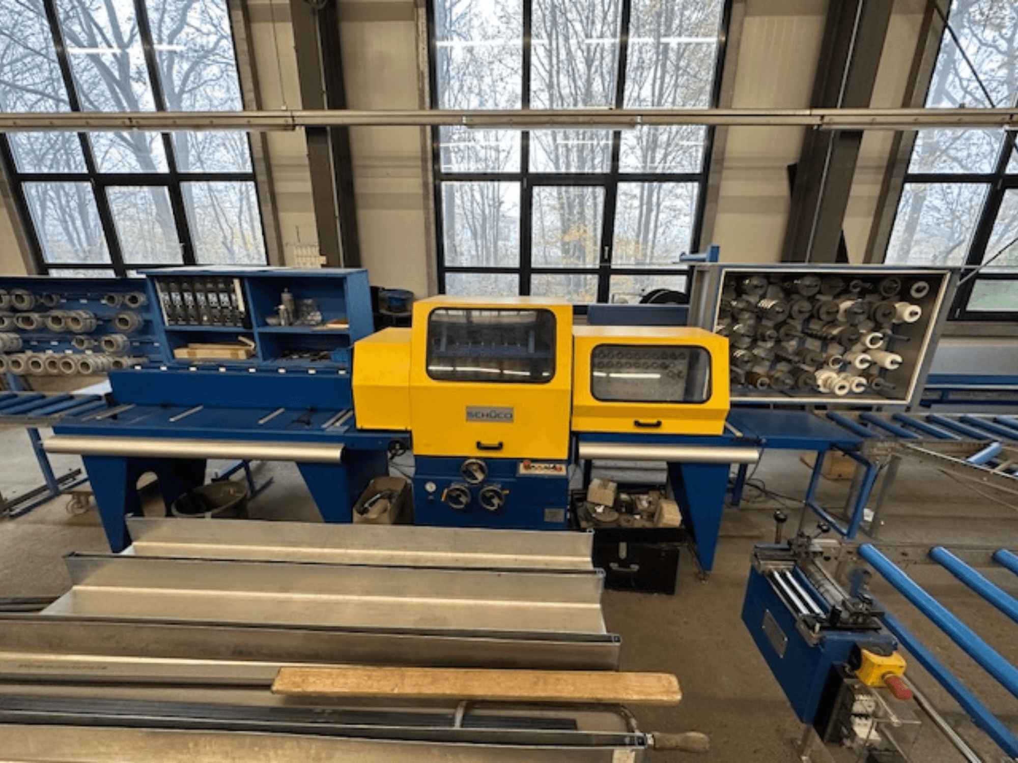 Etukuva SCHÜCO ISOMAT 4 -työstökoneesta, jossa on keltainen ohjauspaneeli, useita rullia ja säilytyslokero.